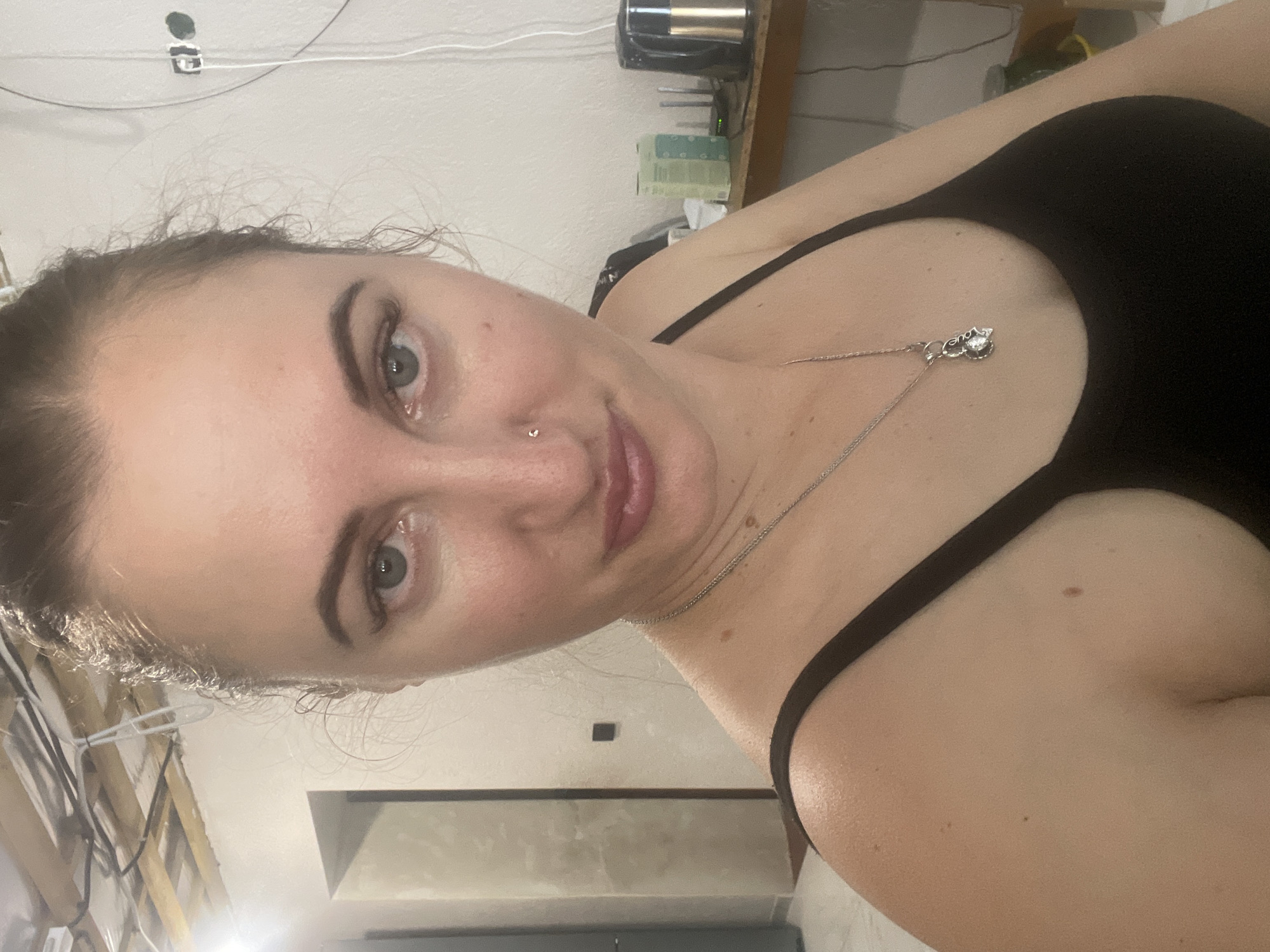 A close-up selfie of a woman in a black tank top, taken indoors. She has light skin, dark eyebrows, and light-colored eyes. She is wearing a silver necklace with a pendant. A nose ring is visible on her right nostril. The background includes exposed ceiling beams and wiring, a white wall with a light switch, and a wooden shelf with some objects on it. The lighting suggests it is artificial light. The location is identified as Zarichne, Ukraine.