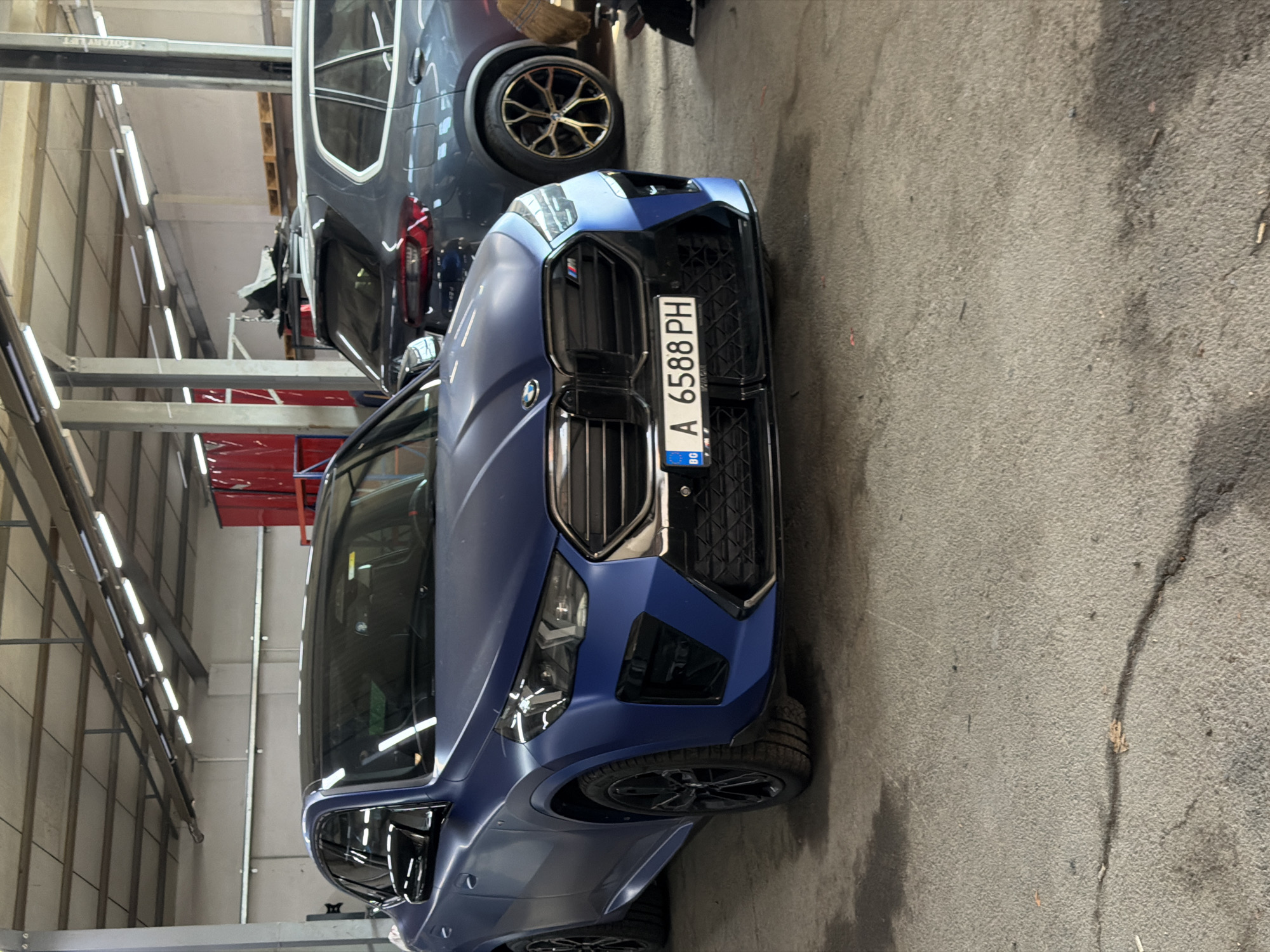 The image displays two cars inside what appears to be an automotive garage or workshop. In the foreground, a matte blue BMW SUV, possibly an X6 or X7 due to its size and design, is parked on a textured concrete floor. The car faces slightly towards the right, showcasing its aggressive front fascia with a large black kidney grille, an M badge, and illuminated headlights. A Bulgarian license plate, "A 6588 PH", is clearly visible on the front bumper. The vehicle is fitted with multi-spoke black wheels.
In the background, a dark gray SUV, partially visible and suspended aloft on a hydraulic car lift, suggests ongoing or impending maintenance. This vehicle also features distinctive multi-spoke wheels with gold/bronze accents. The car lift itself has the text "ROTARY LIFT" visible on its structure.
The environment is an indoor space with bright fluorescent tube lighting running along the ceiling. Metal support beams and light-colored walls are also discernible. The concrete floor shows some wear and cracks. The scene is functional, characteristic of a professional vehicle service center. The visible license plate, with the 'BG' country code and 'A' regional code, confirms the setting is in Bulgaria, consistent with the provided location context of Burgas.