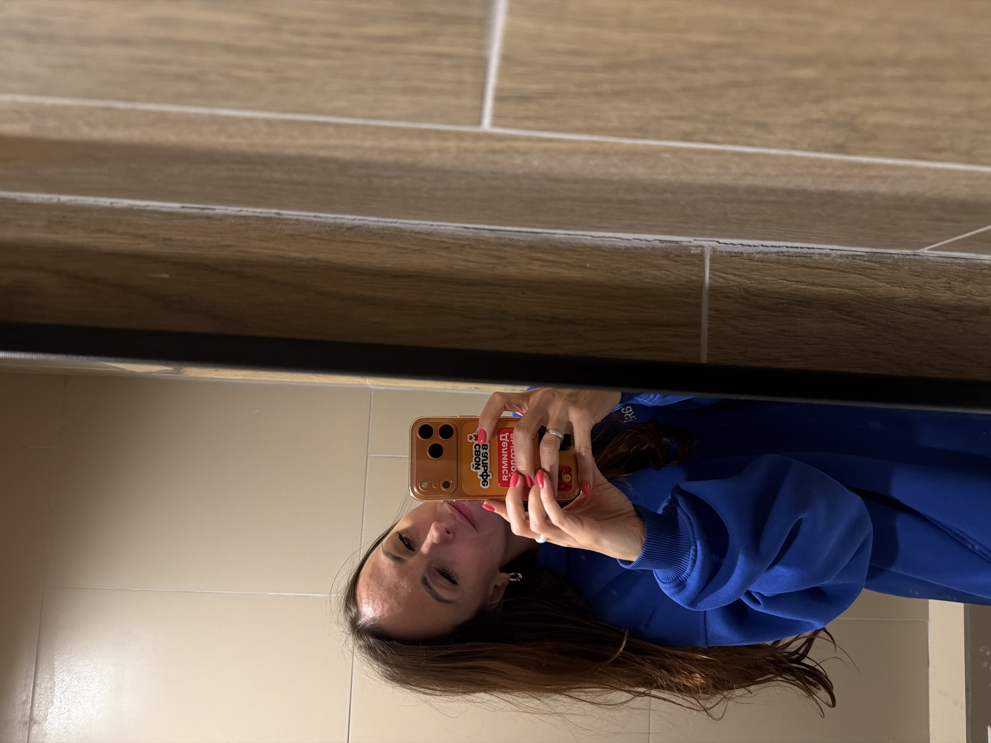 A woman is taking a selfie in a bathroom. She is wearing a blue sweatshirt. The bathroom has tan floor and wall tiles with a wood-grain appearance. The city is not discernible from the image.