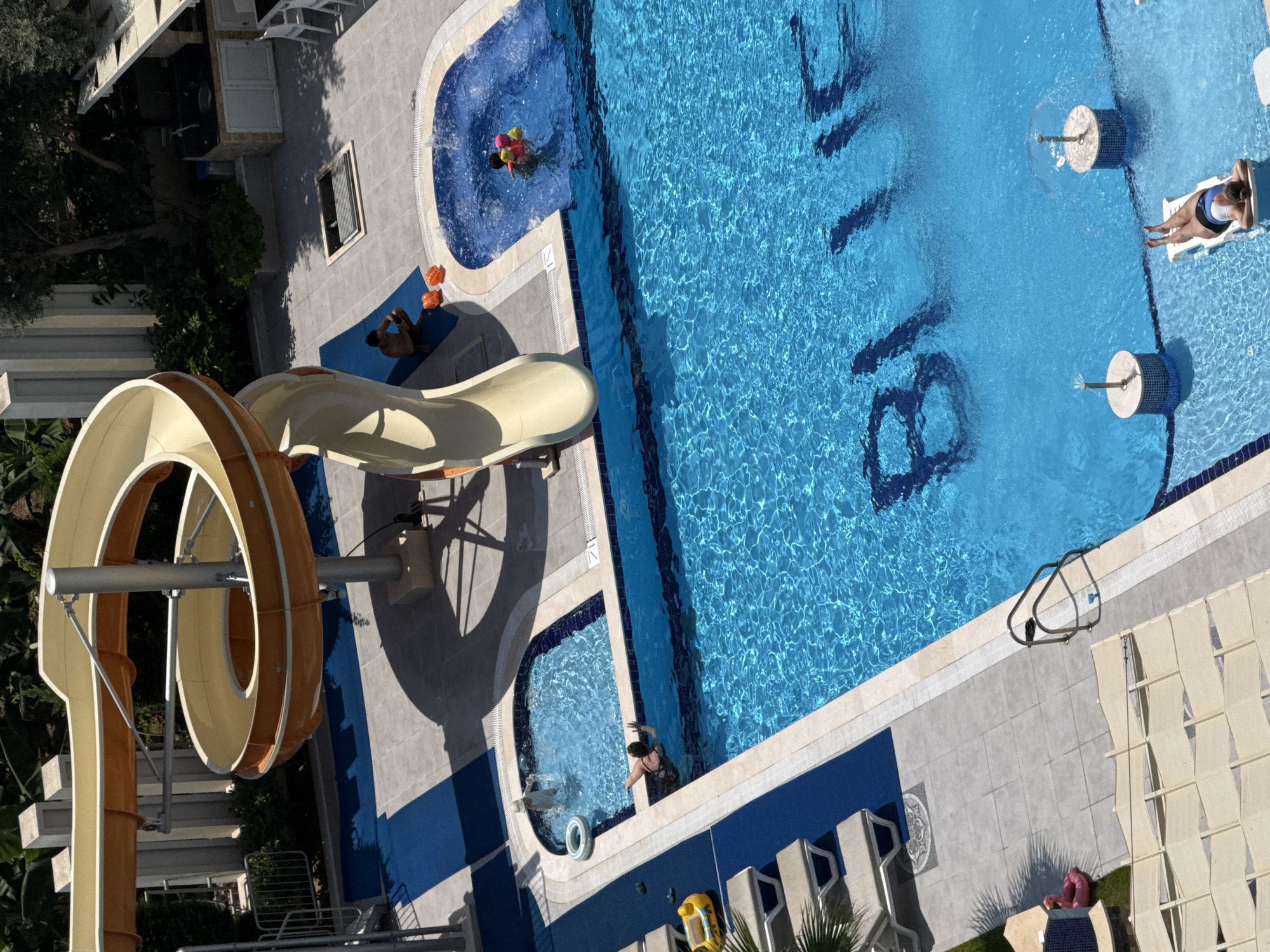 This aerial view captures a resort pool area in Alanya, Turkey, featuring a large, bright blue swimming pool with "BLUE" emblazoned on the bottom. A prominent yellow and orange water slide curves overhead, with its exit landing near a smaller, shallow pool. In the shallow pool, a person in a white head covering is visible. The main pool has a few individuals, including two people in yellow and pink inflatable rings and another person with their arms raised. To the left of the main pool, a person is sitting on a blue mat near the base of the slide, accompanied by two orange objects, possibly toy balls. Further left, lounge chairs are arranged on a tiled patio. A pink inflatable flamingo rests on a patch of grass near the edge of the patio. In the far right of the main pool, a person is relaxing on a white lounge chair partially submerged in the water. Surrounding the pool area are paved walkways, potted plants, and architectural elements typical of a resort, including a covered seating area with tables and chairs in the background. The sun casts strong shadows, indicating a bright, sunny day.