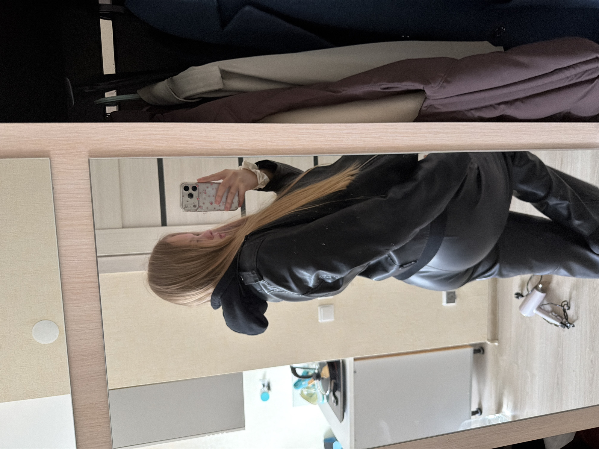 A young, blond woman is seen in the reflection of a mirror, taking a selfie with a floral-printed phone case. She is wearing a black leather jacket and matching pants. The mirror is attached to a dresser or cabinet, above which is a clothing rack with various garments. The background shows the interior of a room, including a light-colored wall and a white cabinet. A hair dryer and other household items can be seen on a nearby surface. There are no discernible details to identify the location as Izhevsk, Russia.