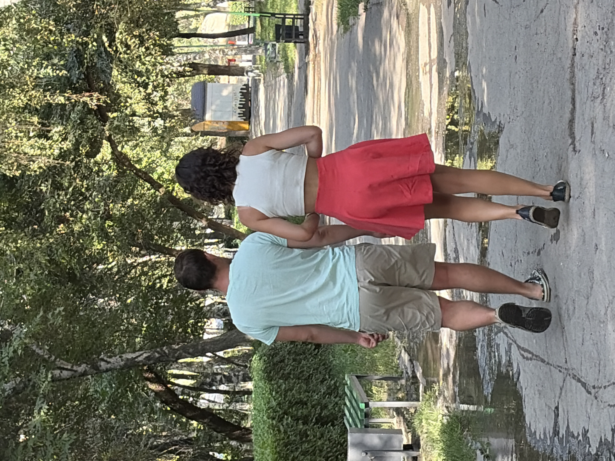 In an outdoor urban park setting, two individuals are walking away from the camera on a paved path. The person on the left is a male wearing a light blue t-shirt and tan shorts. The person on the right is a female wearing a white crop top and a red skirt. The path is wet in places, reflecting the surrounding trees and foliage. To the left of the path, there is a neatly trimmed green hedge and a bench. Further in the background, there are trees with dappled sunlight filtering through their leaves, and several structures, including a white building with a dark roof and some black trash bins and benches. The overall environment suggests a sunny day in a public park.