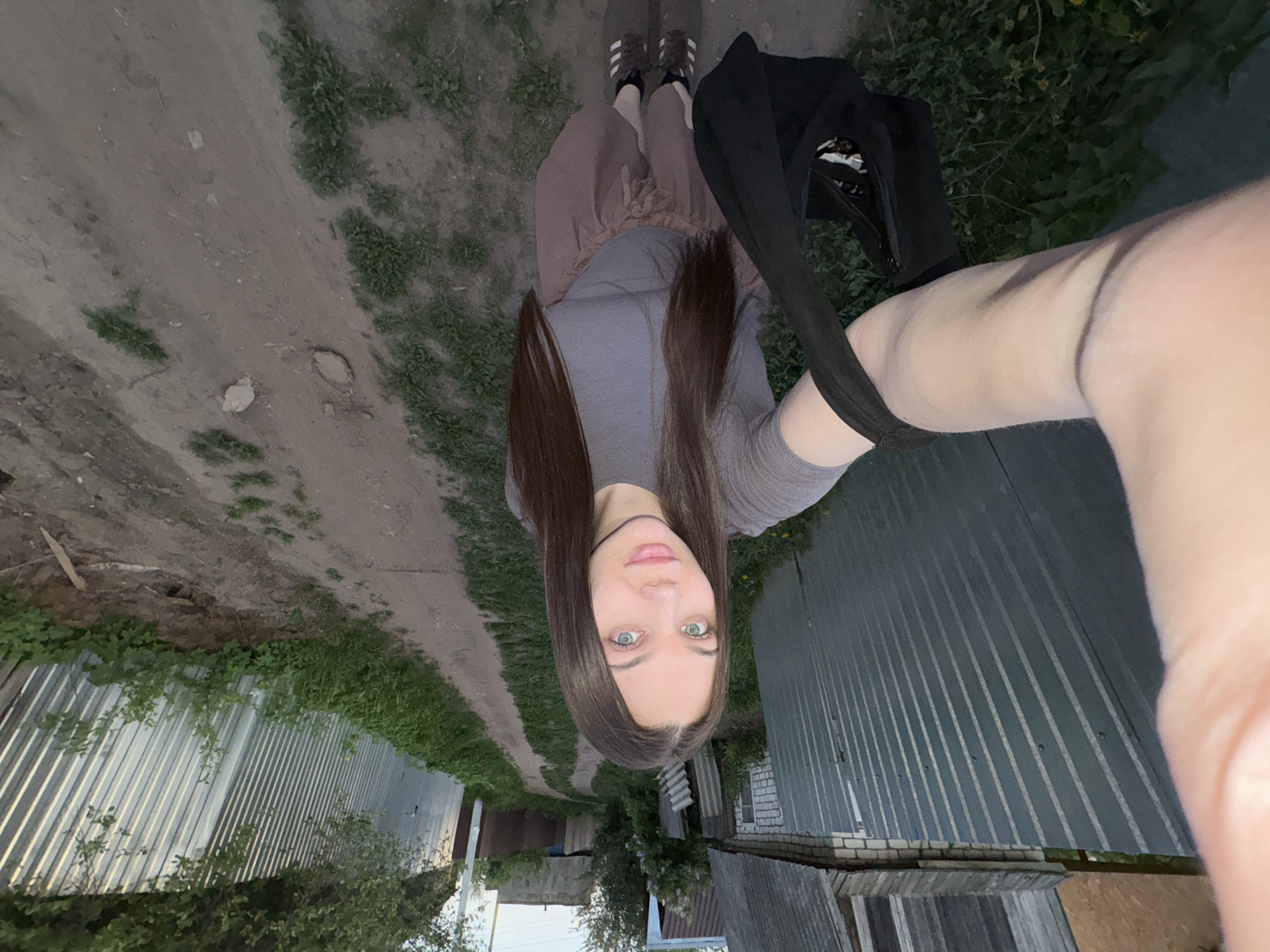 A young woman is taking a selfie in what appears to be an alleyway or dirt road near what appears to be the outskirts of Chaikovsky, Russia. The photo is oriented upside down. She is wearing a gray top and a brown skirt and stands on the dirt road with black sneakers. She has long dark hair and light eyes. She is holding a black bag. On both sides of the dirt road are metal fences, bushes, and vegetation, and the road appears to lead to houses in the distance. The lighting suggests it may be late afternoon or evening.