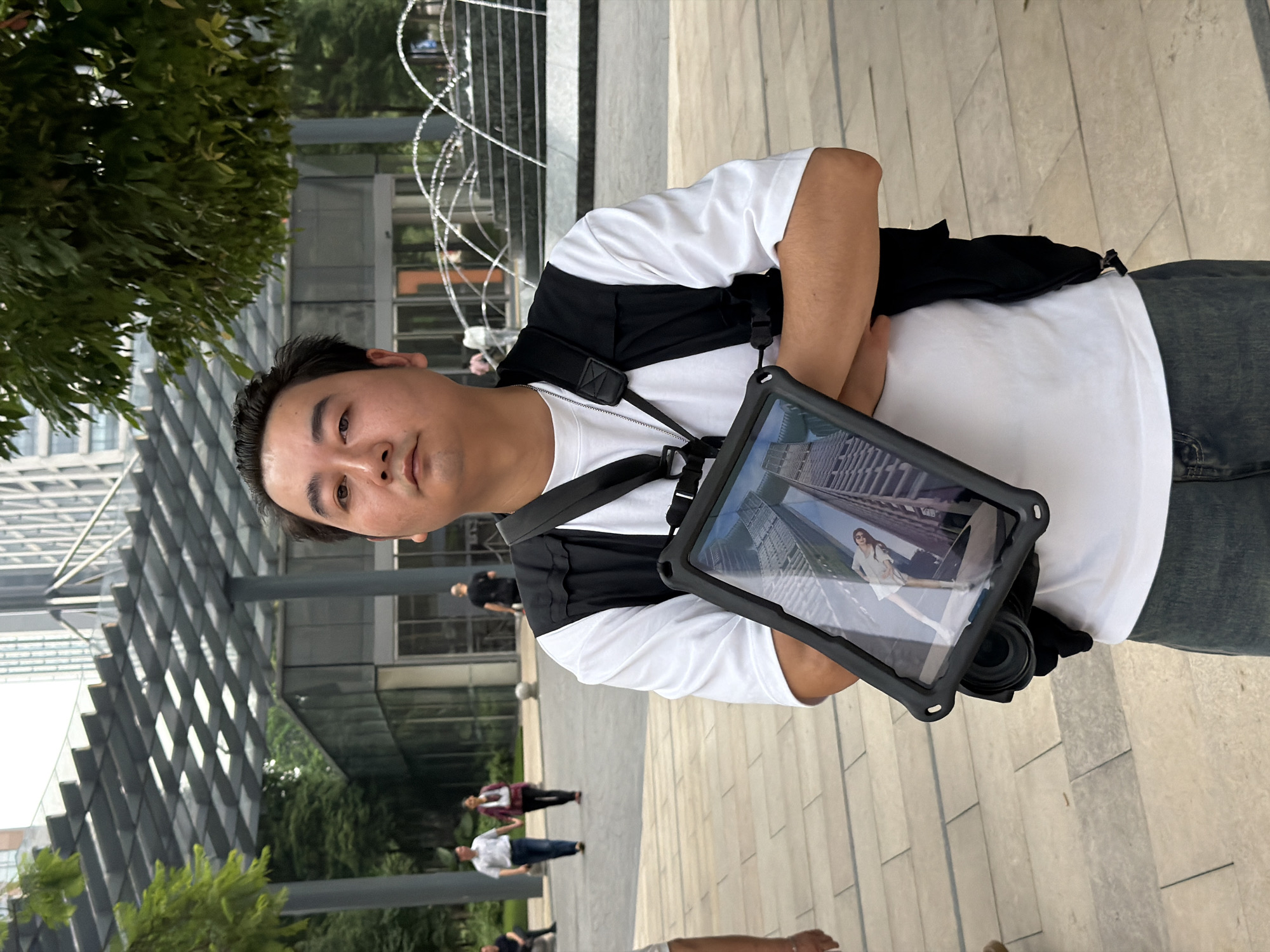 A medium shot shows a man standing outdoors in Yuzhong District, China. The man is wearing a white t-shirt, a black vest, and dark jeans. He has his arms crossed and is looking towards the camera. He is holding a tablet with a camera and what appears to be a lens attached. The tablet displays an image of a woman in a white dress standing in front of a building. In the background, there is a building with a glass facade and a fountain with water jets. Other people are walking in the background. The ground is paved with light-colored tiles.