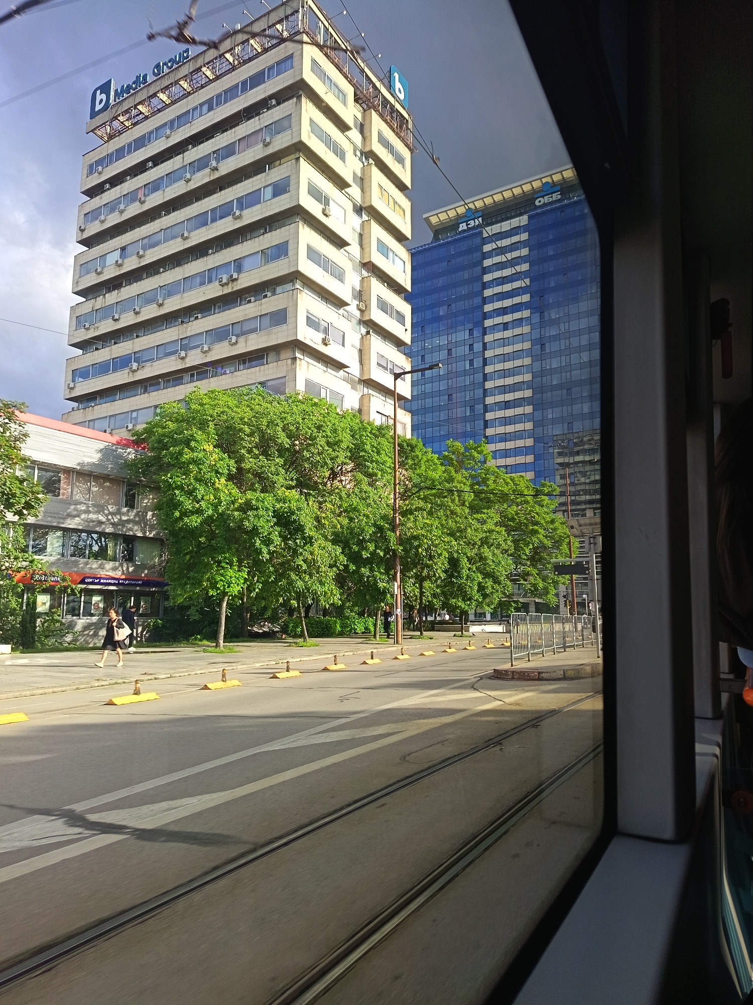 The image captures an urban street scene in Sofia, Bulgaria, viewed from inside a moving tram, with the tram's dark frame visible on the right.
In the foreground, tram tracks run along the street, showing slight motion blur. The asphalt road features white markings and a series of yellow road dividers. To the left, a woman wearing a dark dress and white shoes walks on the sidewalk, carrying a light bag. Behind her, another person is partially visible.
A low-rise building on the left features a "Postbank" sign and the Cyrillic text "ЦЕНТЪР ЖИЛИЩНО КРЕДИТИРАНЕ" (Center for Housing Loans) under a red awning. A row of vibrant green trees lines the street, separating the sidewalk from the main road.
In the background, two prominent buildings dominate the skyline. On the left, a multi-story, light-colored building with numerous windows and air conditioning units features a large blue sign on its rooftop that reads "b Media Group". To its right and slightly behind, a modern, taller skyscraper made of blue-tinted glass stands. Its rooftop also displays Cyrillic text: "ДЗИ" (DZI) and "ОББ" (UBB), indicating DZI and UBB financial institutions.
The scene takes place during the daytime under a partially cloudy sky, with bright sunlight illuminating the buildings and casting shadows on the street, indicating clear weather. The presence of Cyrillic text on buildings confirms the location in Sofia.