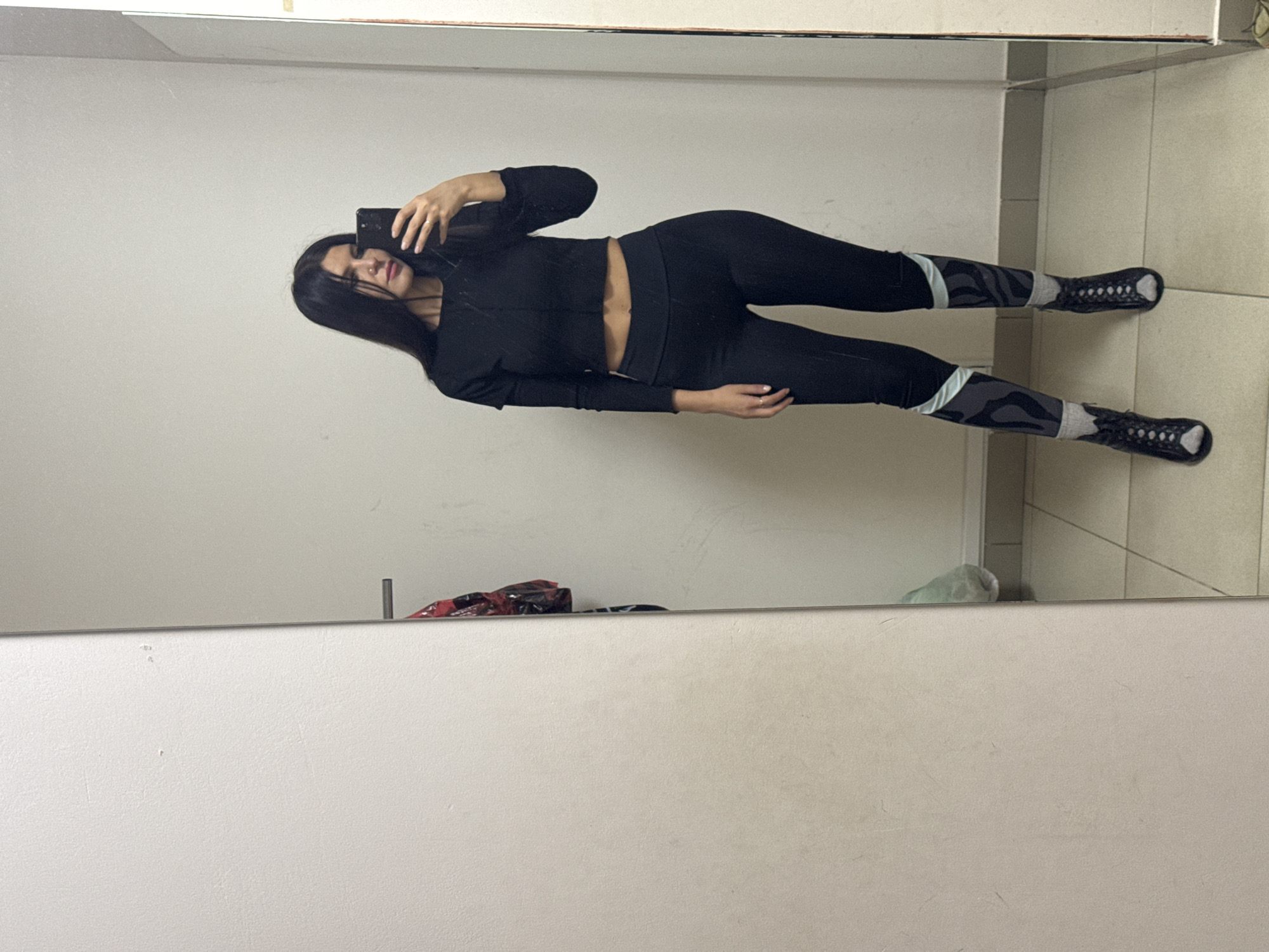 A woman is taking a selfie in a mirror. She has long black hair and is wearing a black long-sleeved crop top, black leggings with grey and black patterns on the lower legs, and black shoes with a lattice-like design. The mirror is in what appears to be a bathroom or changing room, with light-colored tile visible in the upper right corner. Some items, possibly bags, are visible on a surface behind the person in the mirror. It's impossible to determine the exact location in Chaikovsky, Russia, without further information.