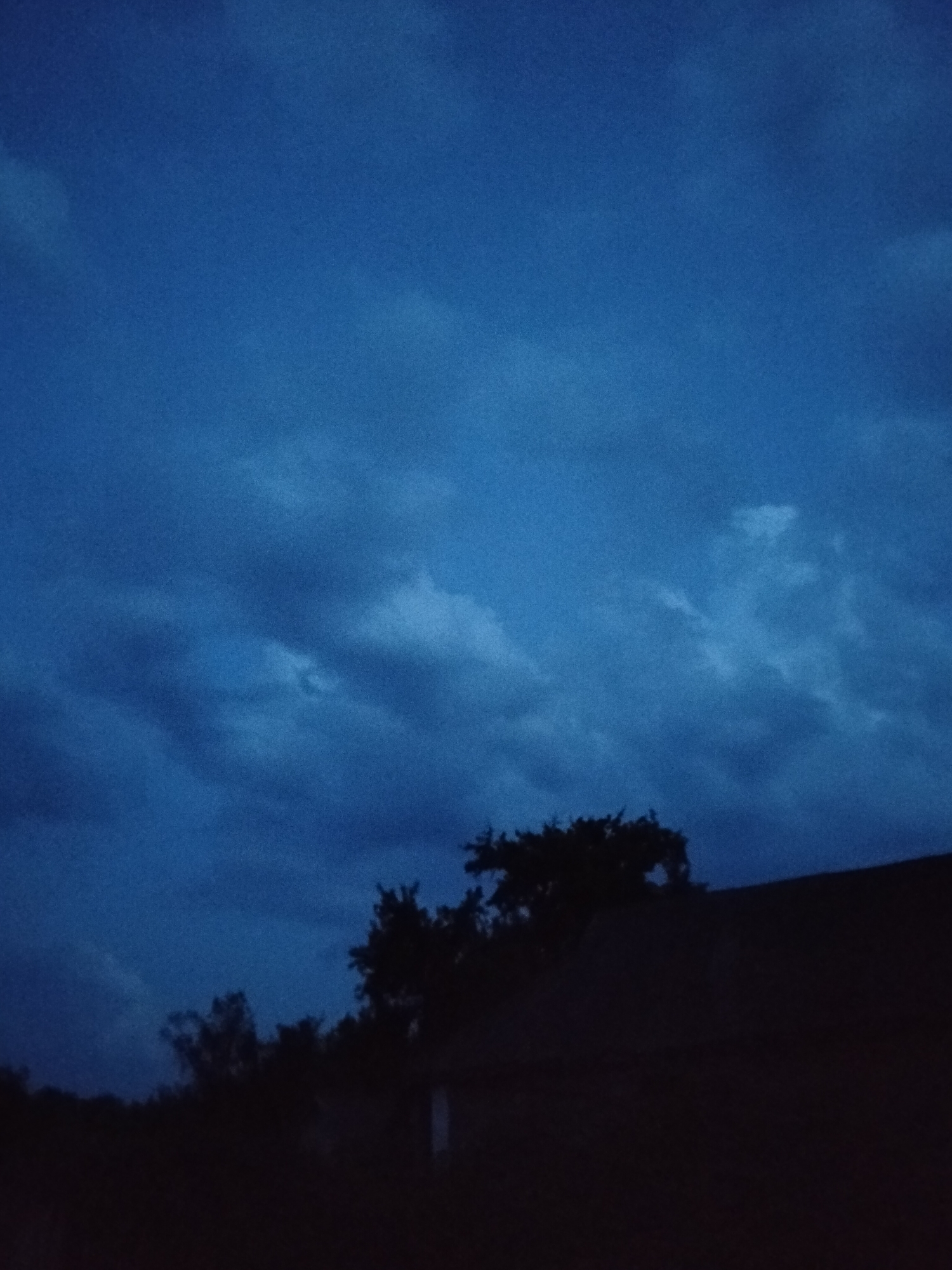 A dark, moody image captures the twilight sky above Ustyvytsia, Ukraine. The sky is a deep, rich blue, with scattered clouds creating a textured canvas. In the foreground, the silhouette of a dark structure, possibly a house with a sloped roof, is visible. Trees, also in silhouette, are scattered to the left and right of the structure, their dark forms contrasting with the slightly lighter sky. The overall impression is one of a quiet, fading evening.