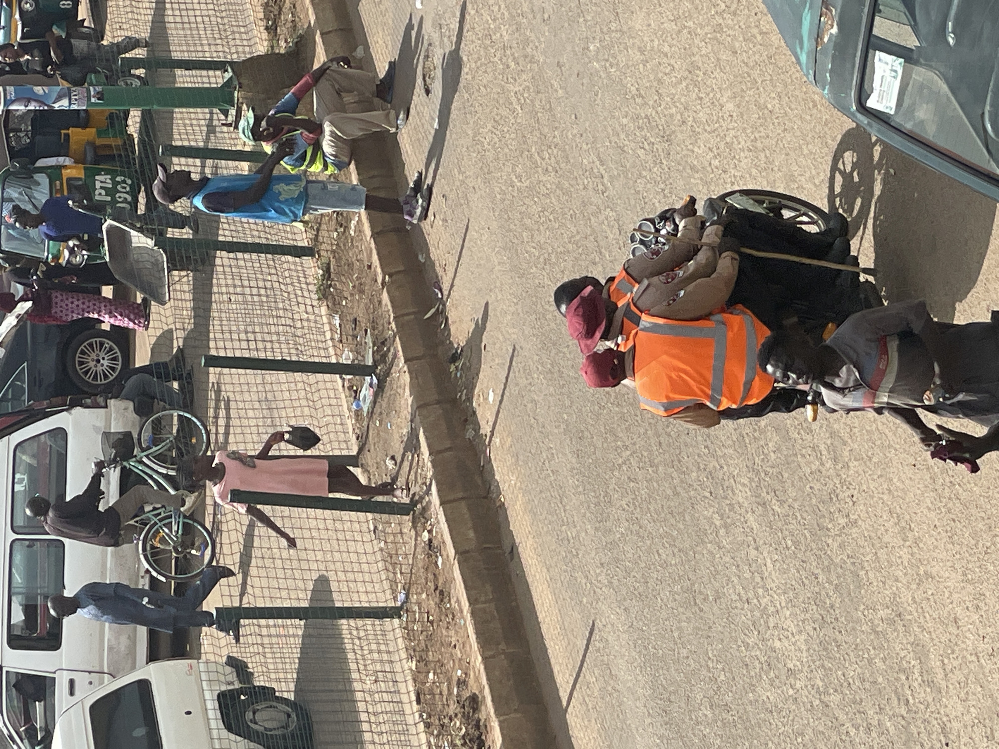 In the city of Unknown, Unknown, on a sun-drenched street, a group of people are captured in a moment of daily life. A young girl in a pink dress walks past a wire fence, carrying a dark bag. To her left, a man in a blue outfit walks briskly, while further back, another man rides a bicycle. In the background, a yellow and green motorized rickshaw is parked, with several individuals standing near it. Across the street, two men are seated on a curb; one in a blue tank top and shorts, the other in a high-visibility vest. On the road, three people are on a motorcycle, two wearing bright orange vests. A man in a grey and striped shirt stands near the motorcycle, holding a dark object. A white van and a portion of a dark vehicle are visible on the left and right sides of the frame, respectively, suggesting a busy urban environment. The street is paved with asphalt, and there are shadows cast by the people and vehicles, indicating a bright day. Debris is scattered along the curb and behind the fence, suggesting a lack of consistent waste management.