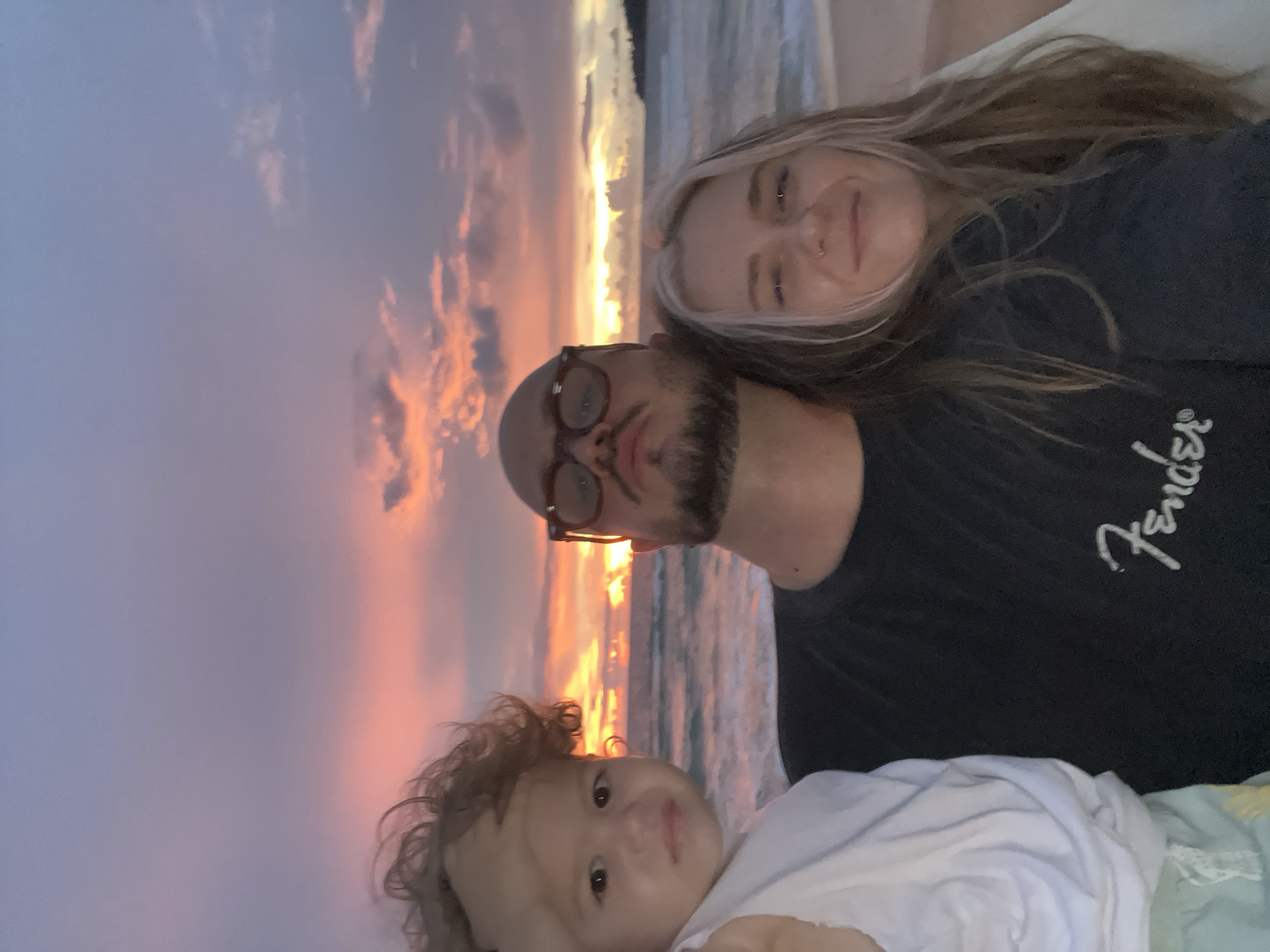 A family of three stands on a beach at sunset. The child in the foreground is being held by a man with a bald head, glasses, and a goatee. The man is wearing a black t-shirt with the Fender logo. To the right of the man, a woman with blonde hair smiles at the camera. The ocean is in the background, with waves gently rolling onto the shore. The sky is a vibrant mix of pink, orange, and purple, with clouds scattered across the horizon. The location is listed as Unknown, Unknown.