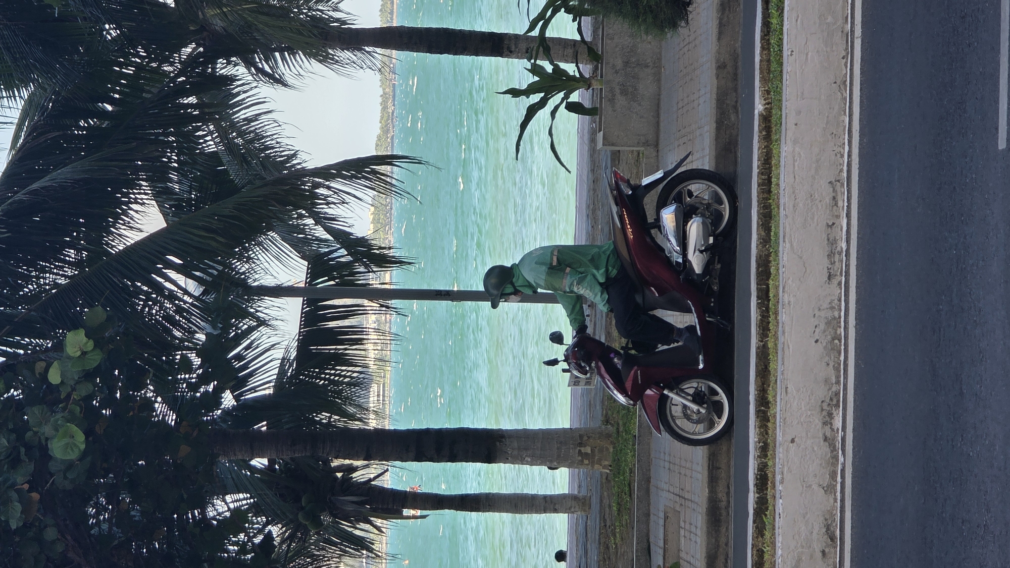 The photo is taken in the city of Bac Nha Trang Ward, Vietnam, and shows a man wearing a green jacket and helmet sitting on a red motorcycle. He appears to be adjusting something on the motorcycle. The motorcycle is parked next to a sidewalk and a road. In the background is a view of the ocean, seen through palm trees. The sky appears hazy.