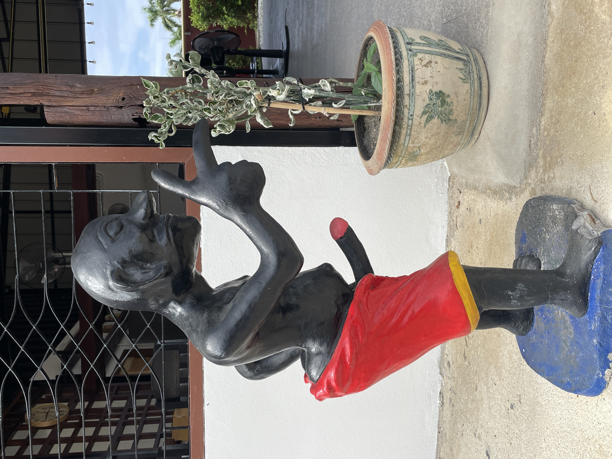The image depicts a black statue of a male figure with exaggerated features, including a prominent nose and a large phallus. The statue is wearing a red loincloth with a yellow trim and stands on a blue base. To the right of the statue is a potted plant with pale green and white variegated leaves, supported by a bamboo stake. The pot itself is ornate, with a light-colored glaze and green decorative motifs. The scene is set outdoors, with a white wall behind the statue and a concrete walkway or patio. In the background, there is a building with wooden posts and a roof, a metal grate over a window, and some palm trees visible in the distance under a cloudy sky. A black electric fan is also visible in the background. The location is specified as Nong Prue, Thailand.