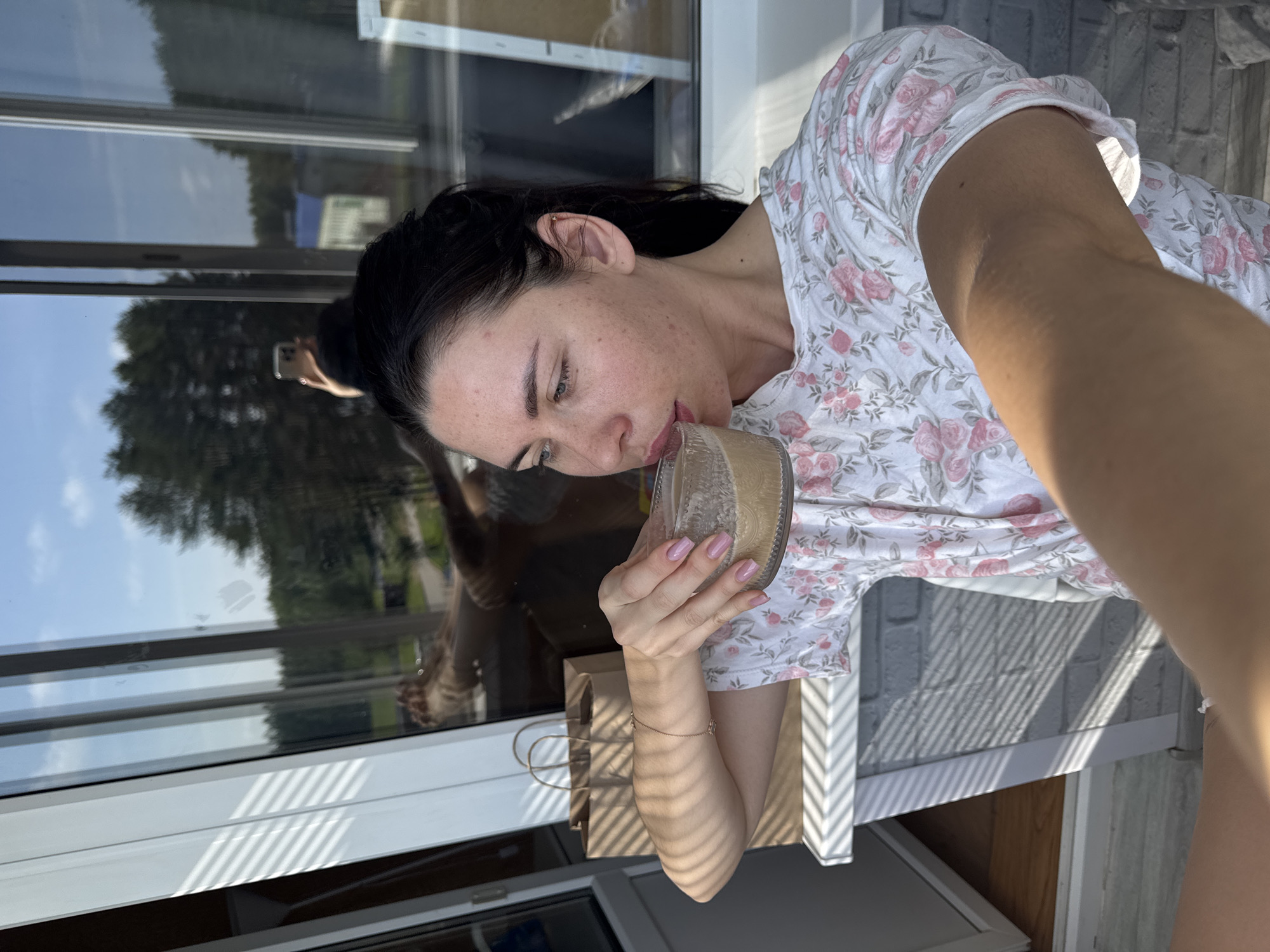 A woman with dark hair pulled back is taking a selfie while drinking from a clear glass. She is wearing a white t-shirt with a floral pattern. In the background, there is a large window showing a wooded area and a blue sky. A brown paper bag is visible to her left. The scene appears to be outdoors on a balcony or patio. The image does not contain any information that places it in the city of Chaikovsky, Russia.