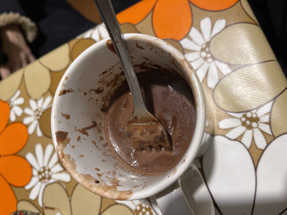 An overhead, close-up shot captures a white ceramic mug, partially consumed, containing a thick, dark brown, chocolate-like substance. A silver metal fork is submerged in the mixture, suggesting either stirring or eating. The inside walls of the mug are streaked and stained with the residue of the chocolate substance. The mug sits on a distinctively patterned surface, likely a tablecloth or placemat, adorned with large, stylized flowers in shades of orange, beige, and white. In the blurred background to the left, a portion of a human hand, possibly wearing a garment with a furry cuff, is visible. The scene suggests an indoor setting, perhaps a casual meal or snack, possibly in a home in Dosso (Ciappanico), Italy, as per the provided context. No text is visible in the image.