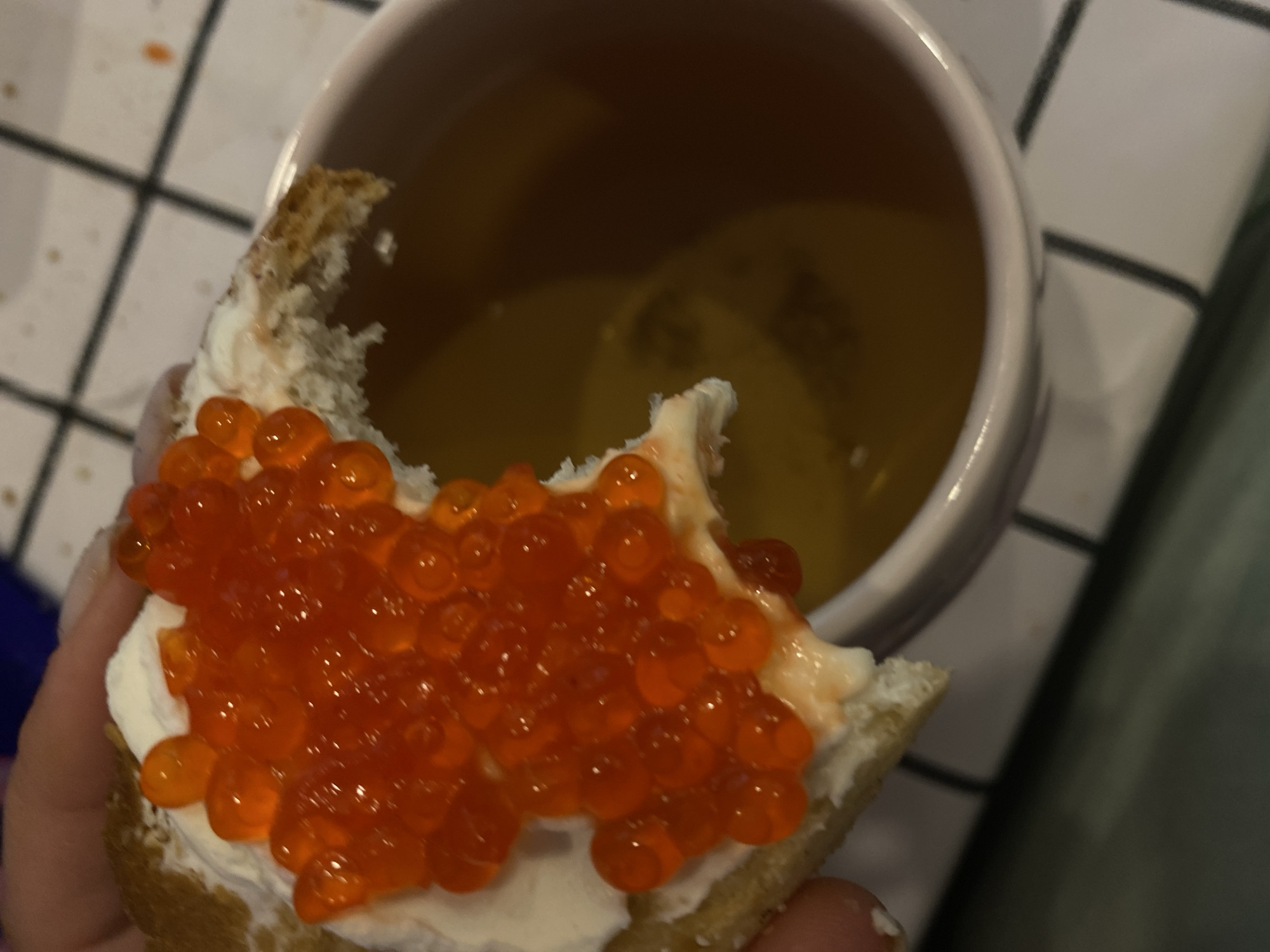 A close-up shot shows a piece of bread topped with cream cheese and red caviar being held by a person's hand, with a pink mug filled with a golden-brown liquid in the background. The bread is triangular and has a bite taken out of it. The caviar is spread generously over the cream cheese, and the individual pearls are visible. The mug is positioned to the right of the bread, and its rim is slightly visible. The background appears to be a checkered surface, possibly a tablecloth or countertop, with the overall lighting suggesting an indoor setting. The image is focused on the food item and the beverage, with no discernible elements that would indicate a specific location in Lomonosov, Russia.