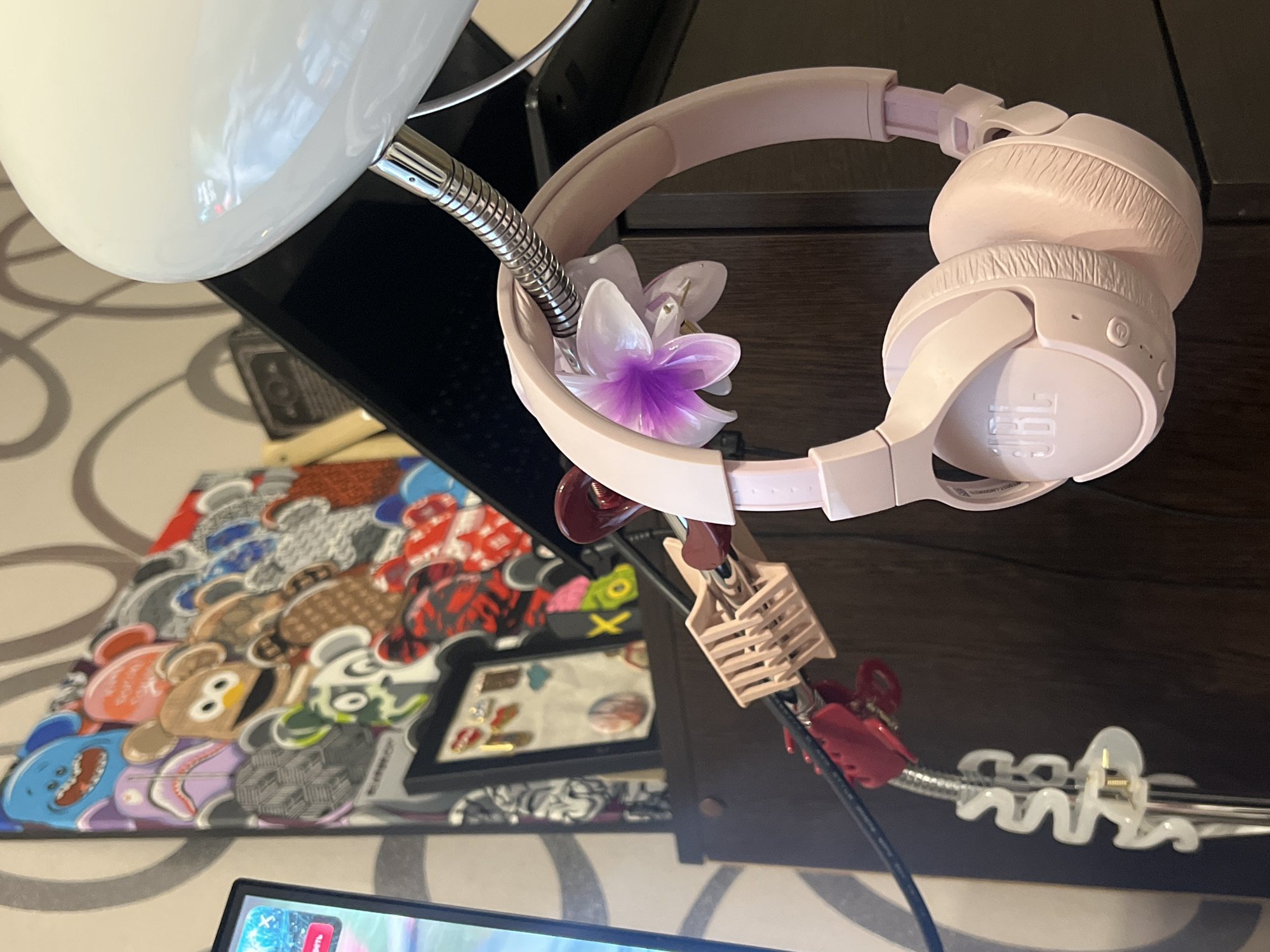 The image displays a close-up, slightly elevated view of a personalized workspace, featuring a light pink JBL over-ear headphone set. The headphones are suspended from a flexible, metallic gooseneck arm, which is part of a glossy white desk lamp partially visible at the top left. A decorative, translucent plastic flower, with white petals fading to a purple center, is tucked between the headphone band and the lamp arm.
Various cable management clips are attached to the gooseneck and cables: a beige ribbed clip, a red claw-style clip securing a black cable, and a white S-shaped organizer. On the dark desk surface beneath, a colorful framed artwork is visible, depicting a collage of stylized, cartoon-like characters or bears in diverse patterns and hues. Behind the cable clips, a smaller black frame holds several pins or badges. A portion of a monitor screen is visible in the bottom left, displaying what appears to be a digital interface with a red button and partially visible text, which includes Russian characters that seem to spell "ОТКРЫТЬ" (Otkryt'), meaning "open." The background features a cream-colored wall with a swirling pattern. The scene is static, highlighting an organized indoor environment, potentially in Kazan, Russia, given the visible Russian text.