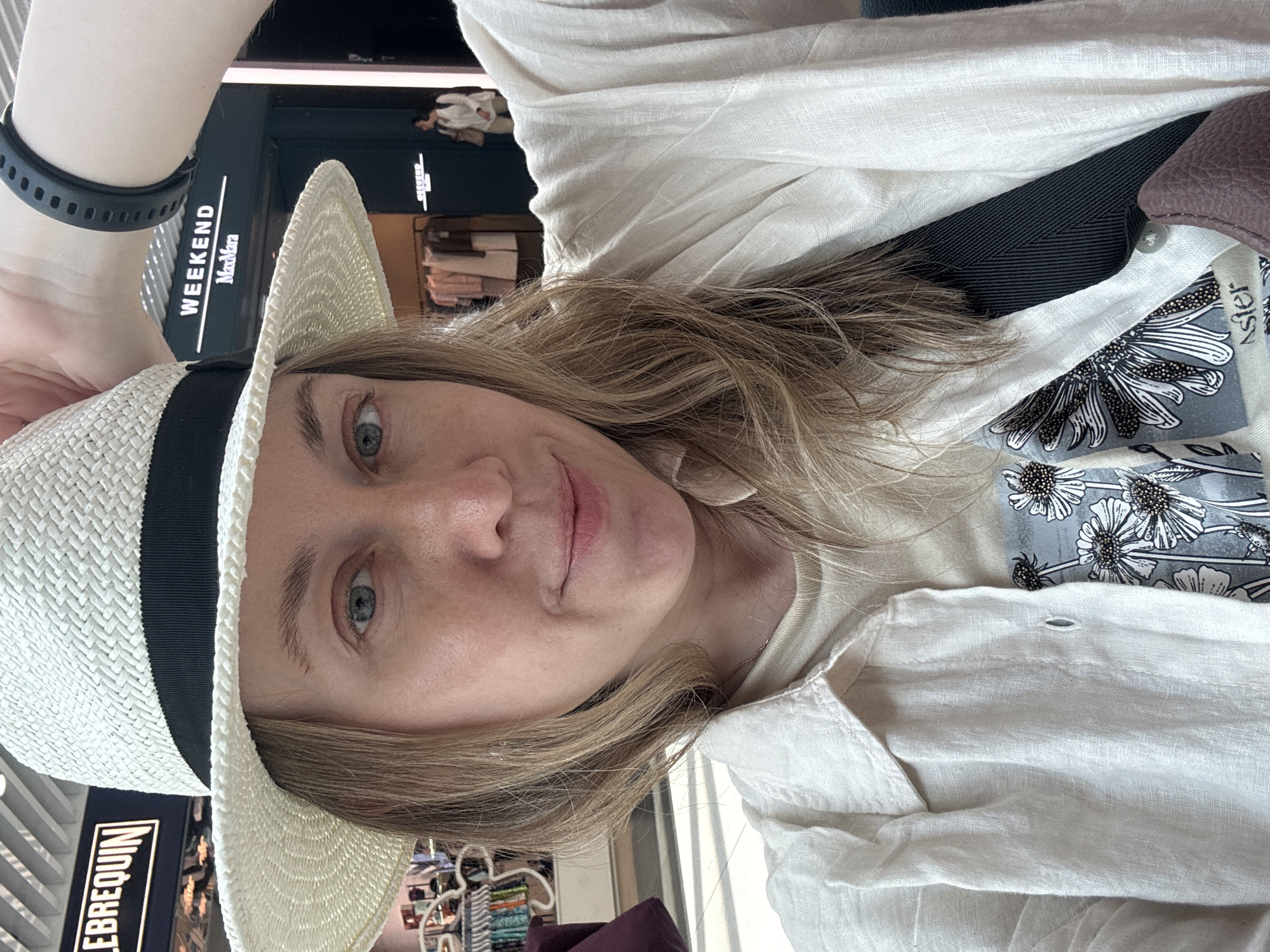 A blonde woman is taking a selfie in Fiumicino, Italy. She is wearing a white straw hat with a black band, a light-colored linen shirt, and a light-colored t-shirt with a floral print. She has a black smartwatch on her left wrist. She is holding the brim of her hat with her right hand. The background shows a store with the words "WEEKEND MaxMara" and another store with the word "LEBREQUIN" visible. A few other people are in the background, and clothing racks are visible in the stores. The lighting suggests it is daytime.