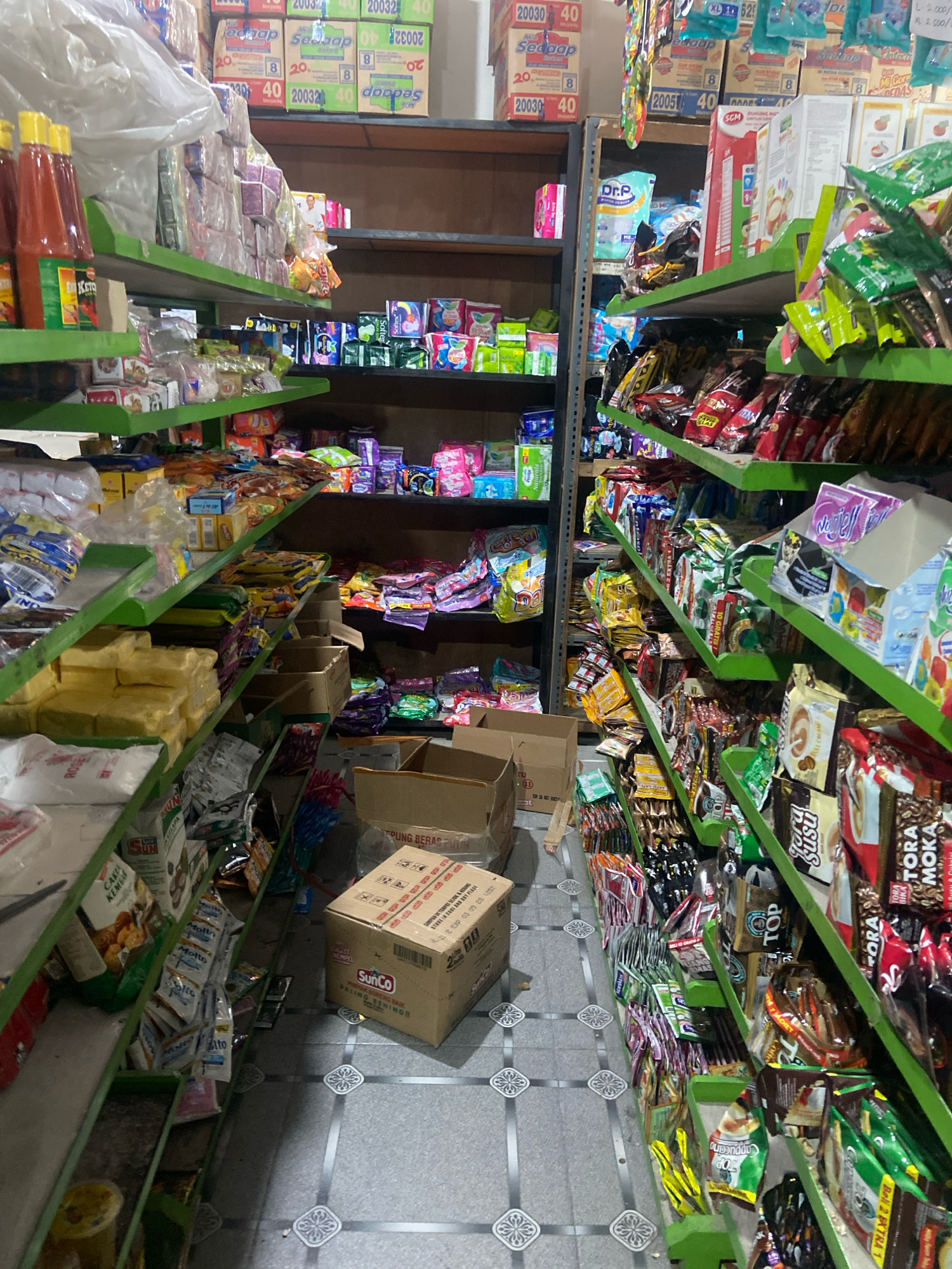 The image depicts the interior of a small convenience store, likely a warung or minimarket, situated in Kaliwungu, Indonesia. The store is characterized by its narrow aisles and densely packed shelves filled with a variety of consumer goods. The shelves are painted a distinctive bright green and are organized into multiple tiers, displaying a wide assortment of packaged food items, snacks, beverages, and personal care products.
Prominently visible on the shelves are numerous brightly colored snack packages, including bags of chips and candies. Also present are boxes of instant noodles, sachets of coffee and tea, and bottles of condiments like ketchup. Larger items such as large sacks of rice or flour are stacked on lower shelves and on the floor. In the foreground, several cardboard boxes are visible, some appearing to contain products for sale, like a SunCo branded box. The floor is tiled with a patterned design common in Indonesian households and shops.
The lighting in the store appears to be artificial, casting a somewhat dim glow on the products. The overall impression is that of a well-stocked, albeit compact, retail space designed to cater to the daily needs of the local community. The presence of Indonesian language on product packaging confirms the location.