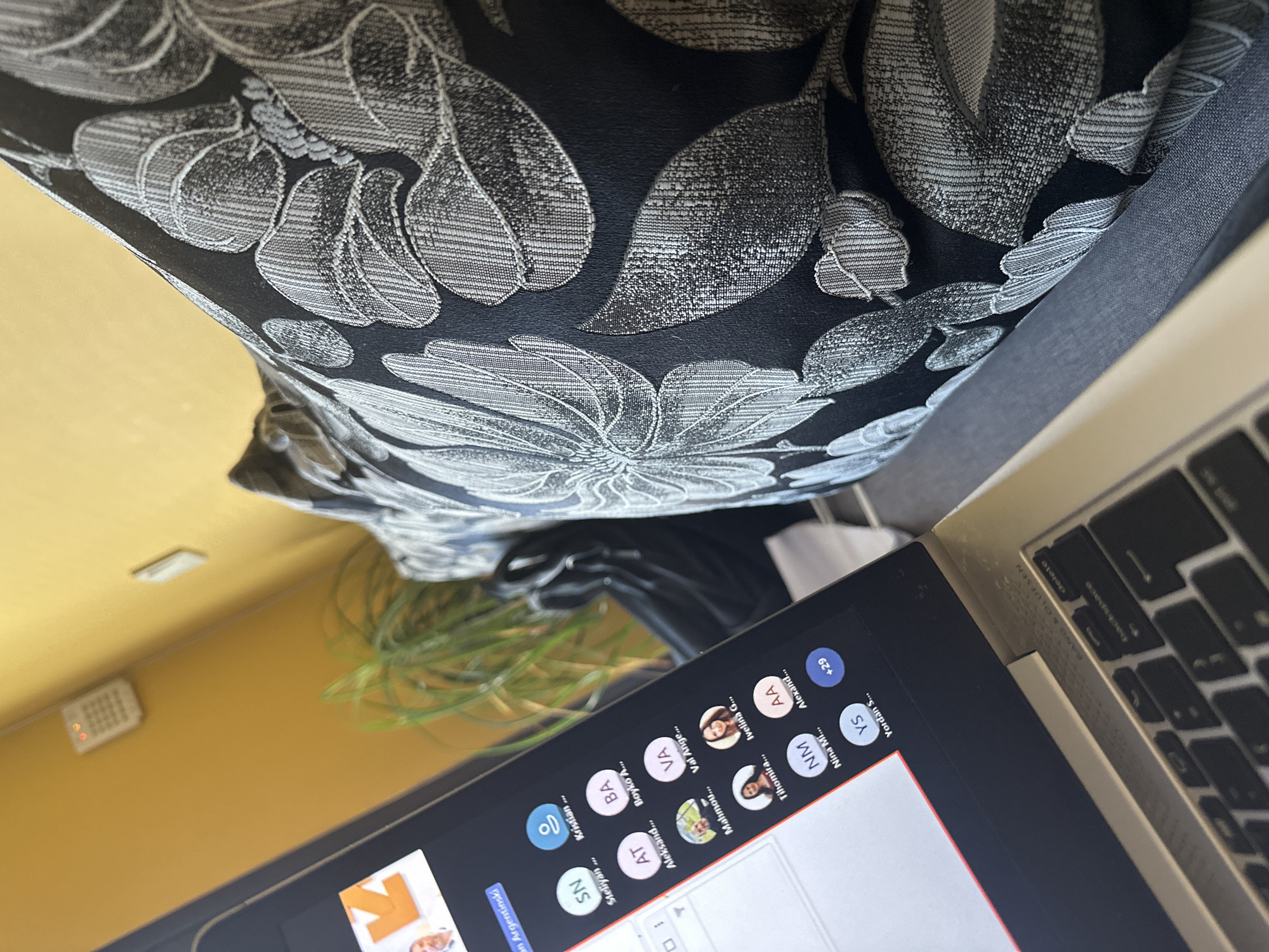 The image shows a laptop screen with a video conference call in progress. Multiple participants are visible in the call, represented by profile icons or initial letters. The laptop sits on a surface partially covered by a floral patterned fabric. In the background, a yellowish wall and a plant are visible. A security keypad is mounted on the wall. Based on the presence of the "Yordan S..." in the list of participants, it is plausible that the image might originate from the city of Sofia, Bulgaria.