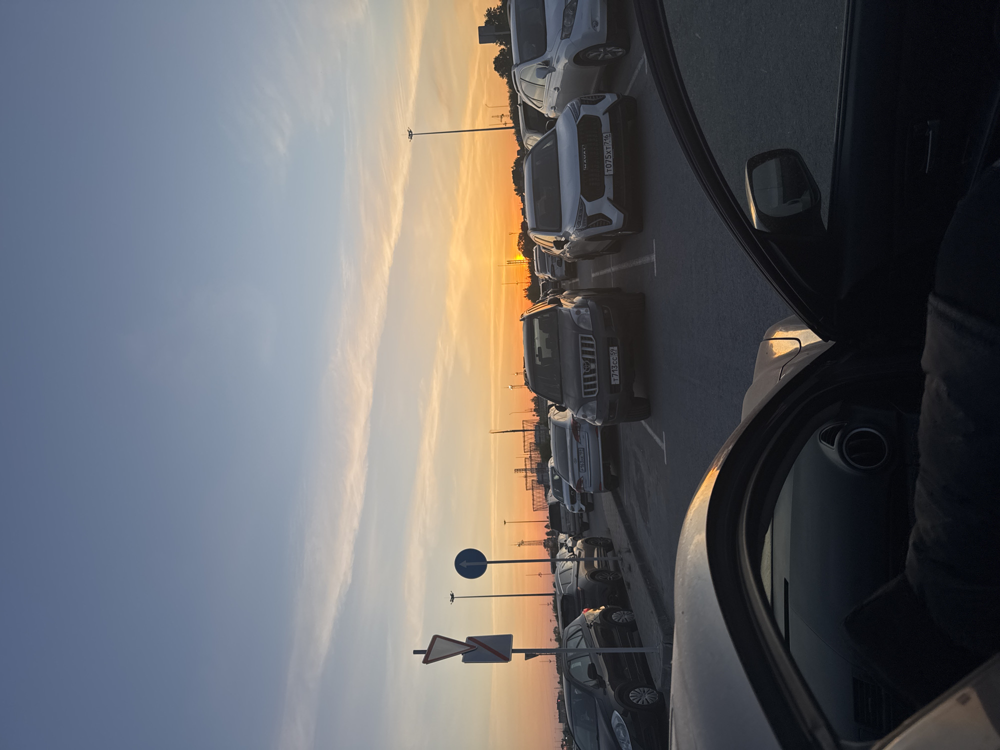 The image depicts a parking lot in Bolshoye Savino, Russia, at sunset. A row of cars is parked diagonally along the right side of the frame, with the open door of a car on the right side dominating the foreground. The sunset casts an orange glow in the background, between cars and poles. Road signs are visible in the middle of the frame. The sky is blue near the top.