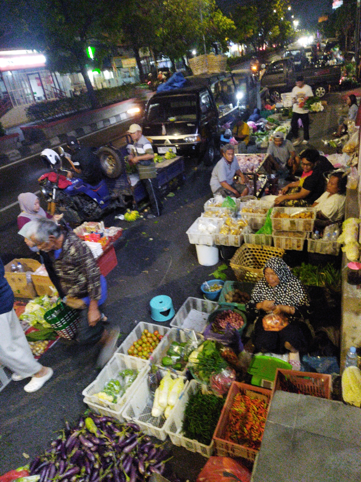 An outdoor marketplace scene is depicted at night, likely in Semarang, Indonesia, with various vendors and shoppers interacting amidst a variety of produce and goods. The overall lighting is dim, with artificial lights contributing to the visibility.
In the foreground, a large pile of purple eggplants is spread across the ground. To the right of the eggplants, a red plastic crate is filled with red and orange chili peppers. Behind these, a woman wearing a black and white patterned headscarf and a dark garment is seated, holding a plastic bag of goods and looking down. She is positioned next to a gray textured surface, possibly a wall or counter.
Further to the left, white plastic crates are stacked and filled with produce. One crate contains small red and green tomatoes. Another holds green vegetables, possibly bell peppers. A third crate contains yellow items, possibly corn or banana blossoms. There are also woven baskets, one of which is large and cylindrical and filled with a dark material.
In the mid-ground, a man in a patterned shirt and blue pants is walking, holding a basket of green produce. He appears to be a vendor. Near him, a motorcycle with a blue seat and a white helmet on it is parked. A black car is partially visible, and a person in a white shirt and dark pants is standing near it, holding a bunch of yellow fruit.
In the background, trees with dark foliage line the street. Buildings with illuminated windows are visible. Several other vehicles, including cars and possibly scooters, are present. A few individuals are seen walking or standing in various parts of the scene. The ground is paved with asphalt. The atmosphere suggests a bustling night market.