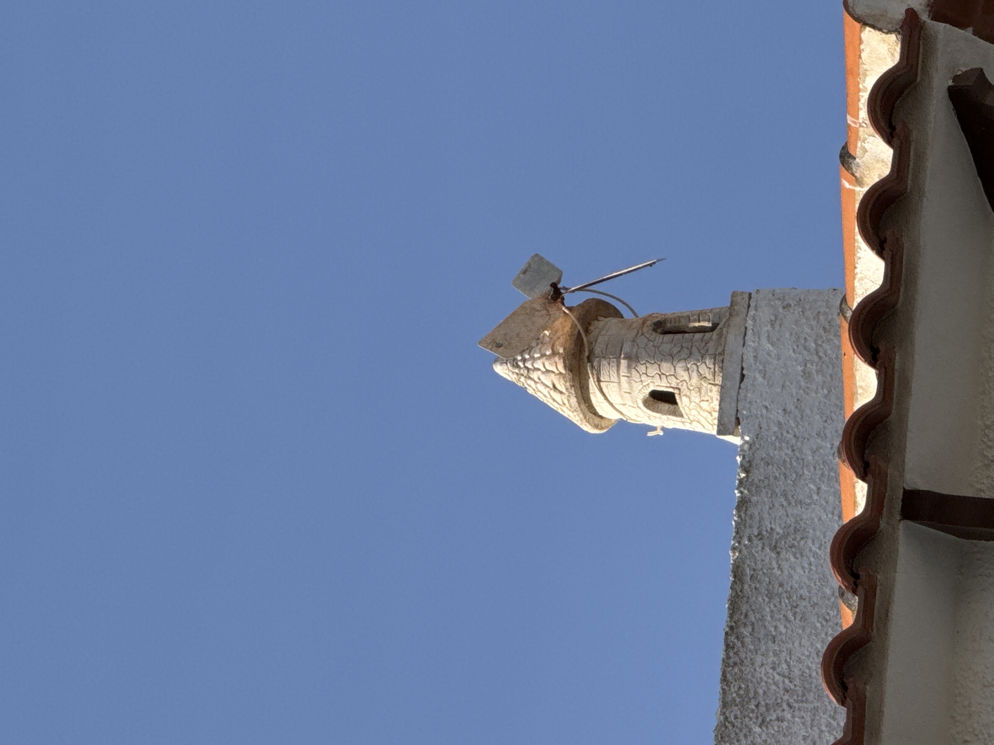 The image captures a close-up view of a decorative windmill chimney atop a white textured wall, set against a clear, bright blue sky. The windmill is a miniature replica, constructed from a light-colored stone-like material, with a conical roof and a small arched window. A simple, two-bladed propeller extends from the top. Below the windmill, the textured white wall extends to the left, and the edge of a red-tiled roof is visible in the foreground, obscuring the lower portion of the structure. The overall scene suggests a building or architectural feature in an outdoor setting, likely in a Mediterranean climate, such as Torrevieja, Spain, known for its distinctive architecture and coastal environment.