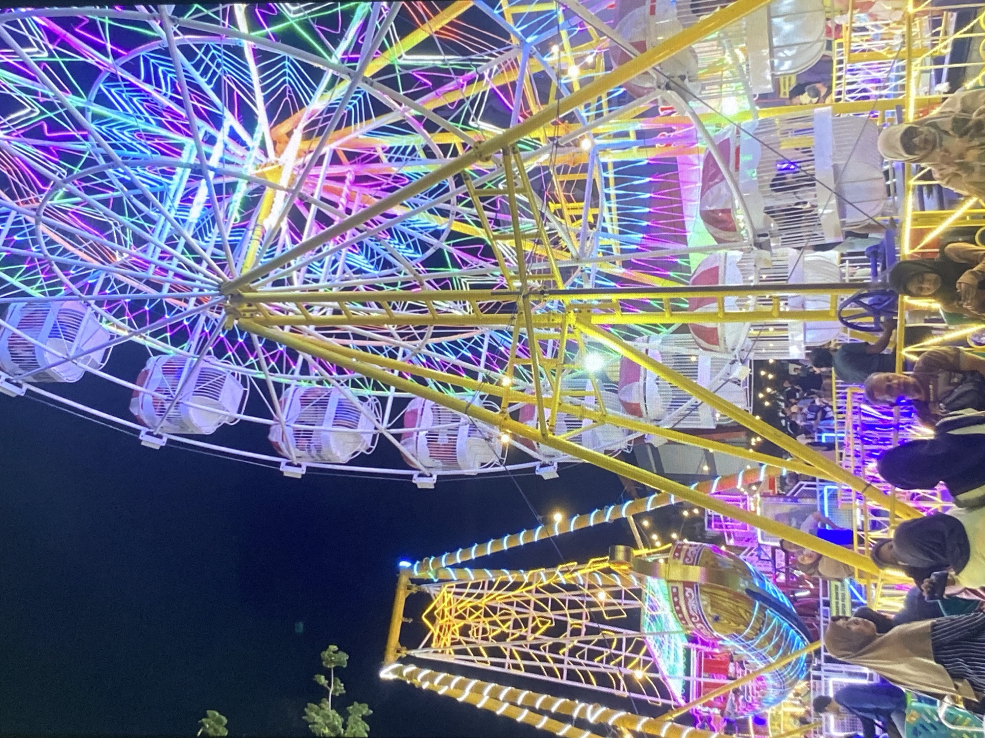 This image depicts a vibrant night scene at an amusement park in Tebel, Indonesia, likely during a festival or event. Dominating the frame is a large Ferris wheel adorned with a dazzling array of colorful LED lights in shades of pink, purple, blue, and green, creating a geometric and radiant pattern against the dark night sky. To the left, a pirate ship-themed swing ride, also illuminated with yellow and blue lights, stands tall.
In the foreground and mid-ground, several people are visible, casually dressed and seemingly enjoying the atmosphere. Some are looking at their phones, while others are conversing. A group of women wearing hijabs are prominent in the lower part of the image, with one woman in a beige hijab and a striped top looking towards the camera. The environment suggests a lively public gathering, with the bright lights of the rides creating a festive ambiance. The overall scene captures the excitement and visual spectacle of a night at the amusement park.