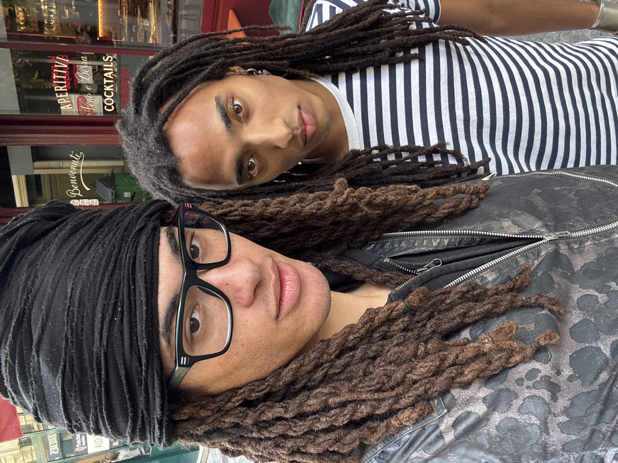 The image, rotated 90 degrees clockwise, presents a close-up portrait of two individuals with dark dreadlocks, likely a selfie, with a storefront visible in the background.
The individual in the foreground is partially visible, wearing a black headwrap or bandana that covers part of their head. They are wearing black-framed glasses and have dark brown dreadlocks that frame their face and extend over their shoulders. A hint of stubble is visible on their chin. Their expression appears neutral, with their lips slightly parted. They are wearing a black jacket with a prominent zipper and a subtle, dark leopard or abstract spotted pattern.
Behind and slightly above them is another individual, whose face is more fully visible. They have dark brown dreadlocks cascading around their head and shoulders. They are wearing a white t-shirt with thin black vertical stripes. A silver earring is visible in their left ear. They have dark eyes and are looking directly at the camera with a direct, slightly inquisitive gaze.
In the blurred background, through what appears to be a glass window, a storefront is visible. Red window frames and interior lighting suggest an establishment. Several Italian words are discernible on the glass in red lettering, oriented vertically. "APERITIVO" is prominent, along with "Vino e Birra" (Wine and Beer) and "COCKTAILS". Below "APERITIVO," "Benvenuti" (Welcome) is visible in cursive script. The setting appears to be a street scene in an urban environment, consistent with Paris, France. The lighting suggests it could be evening or an indoor setting.