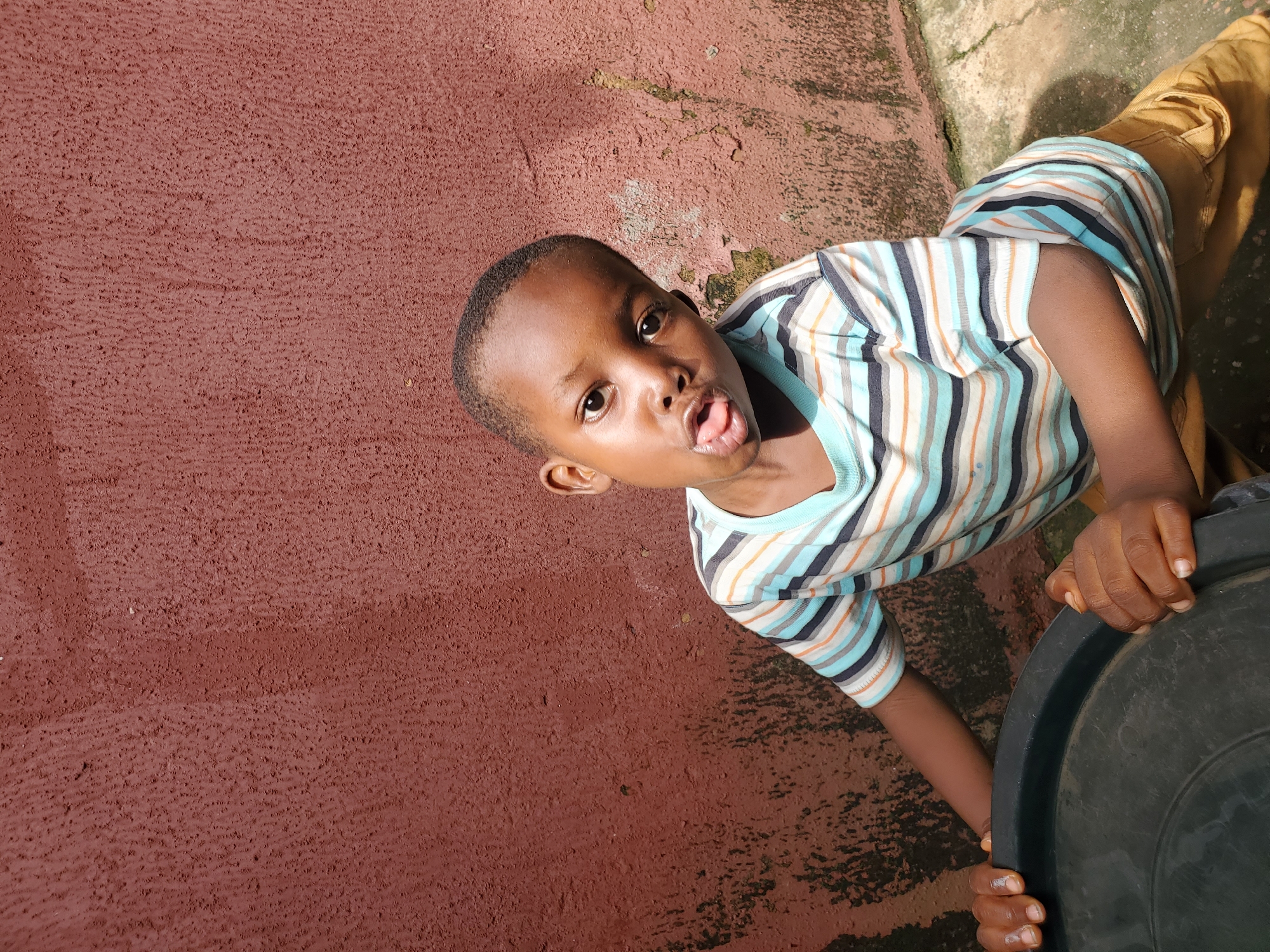 A young Black child, likely a boy, is the subject of this image. He is wearing a striped t-shirt with alternating bands of blue, white, orange, and black, and tan pants. The child is positioned in a way that suggests he is either crawling or in a low crouch, with his face turned towards the camera. He has a playful or mischievous expression, with his tongue sticking out slightly. His left arm is bent, and his hand is resting on a large black plastic object, possibly a bucket or basin. His right arm is also visible, reaching towards the object.
The setting appears to be outdoors. The ground is a textured, reddish-brown surface, possibly concrete or packed earth, with some areas of lighter, more weathered material and dark stains. There are also some shadows visible, indicating sunlight, which suggests it is daytime. The lighting suggests the sun is high in the sky, creating strong highlights and shadows. The overall impression is of a candid moment captured, possibly in a yard or a public outdoor space. The child's expression and the candid nature of the shot convey a sense of youthful energy and playfulness. No text is visible in the image.
