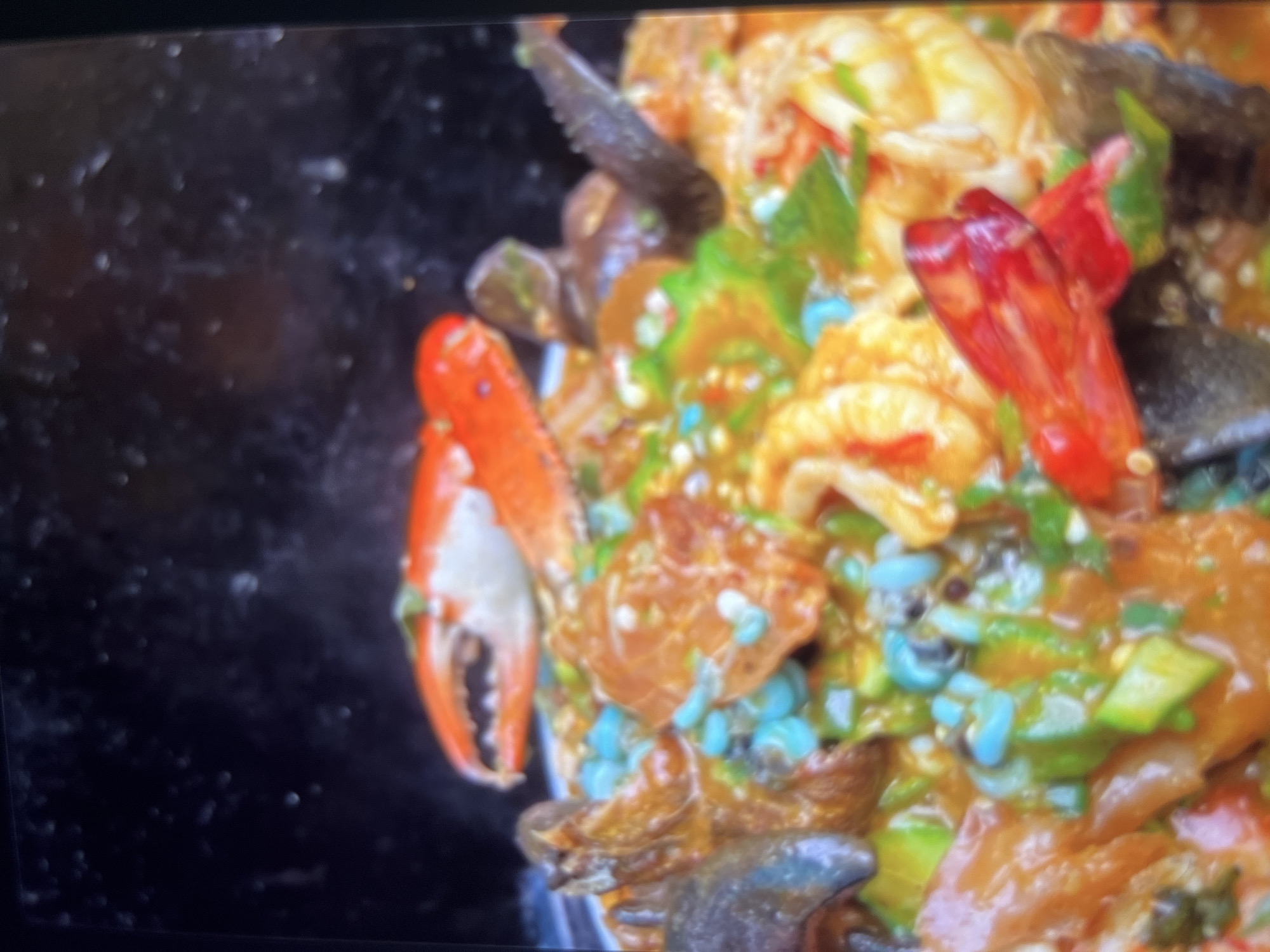 The image shows a close-up of a dish of seafood. The dish appears to be a stew or gumbo, with various types of seafood including shrimp, mussels, and what looks like crab legs. The seafood is in a rich, orange-colored sauce, and there are pieces of green vegetables, possibly okra, mixed in. Some small, bright blue, oval-shaped items are also visible, which might be a type of grain or spice. The dish is presented in a bowl and is very colorful. The background is dark and out of focus, suggesting the image was taken indoors with artificial lighting. There is no discernible information about the location within Abuja, Nigeria.