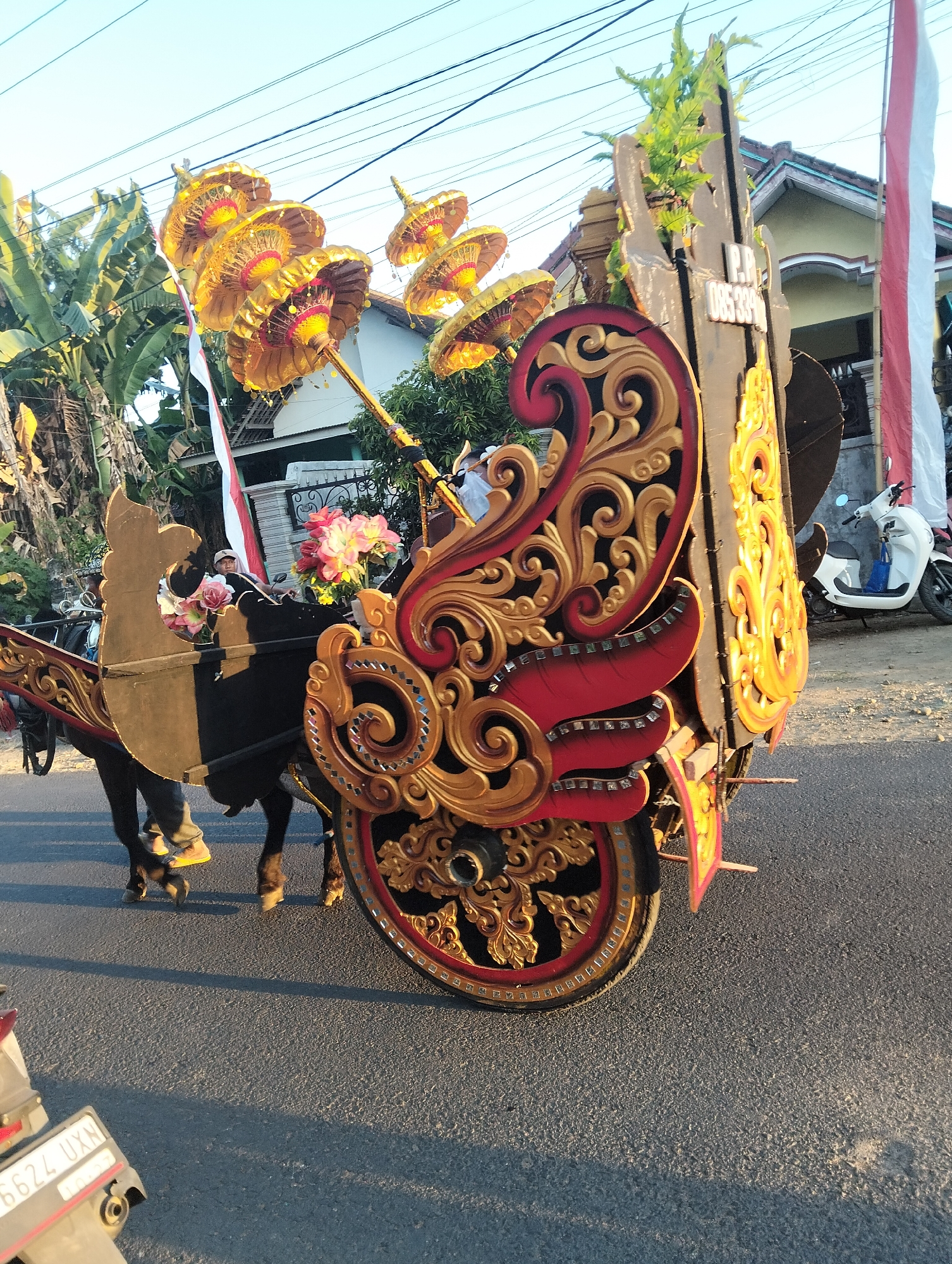 In the city of Sumbersewu, Indonesia, a horse-drawn carriage elaborately decorated with gold and red ornamentation is on a paved road. The carriage features several tiered golden umbrellas with red accents at the top, along with greenery. A person wearing a cap and a light-colored shirt is seated in the carriage, which is adorned with artificial flowers in pink and white. A dark-colored horse is harnessed to the front of the carriage. To the right of the carriage, a white scooter is parked. In the background, a building with a yellow facade and a red-tiled roof is visible, as is an Indonesian flag. Power lines crisscross the sky.