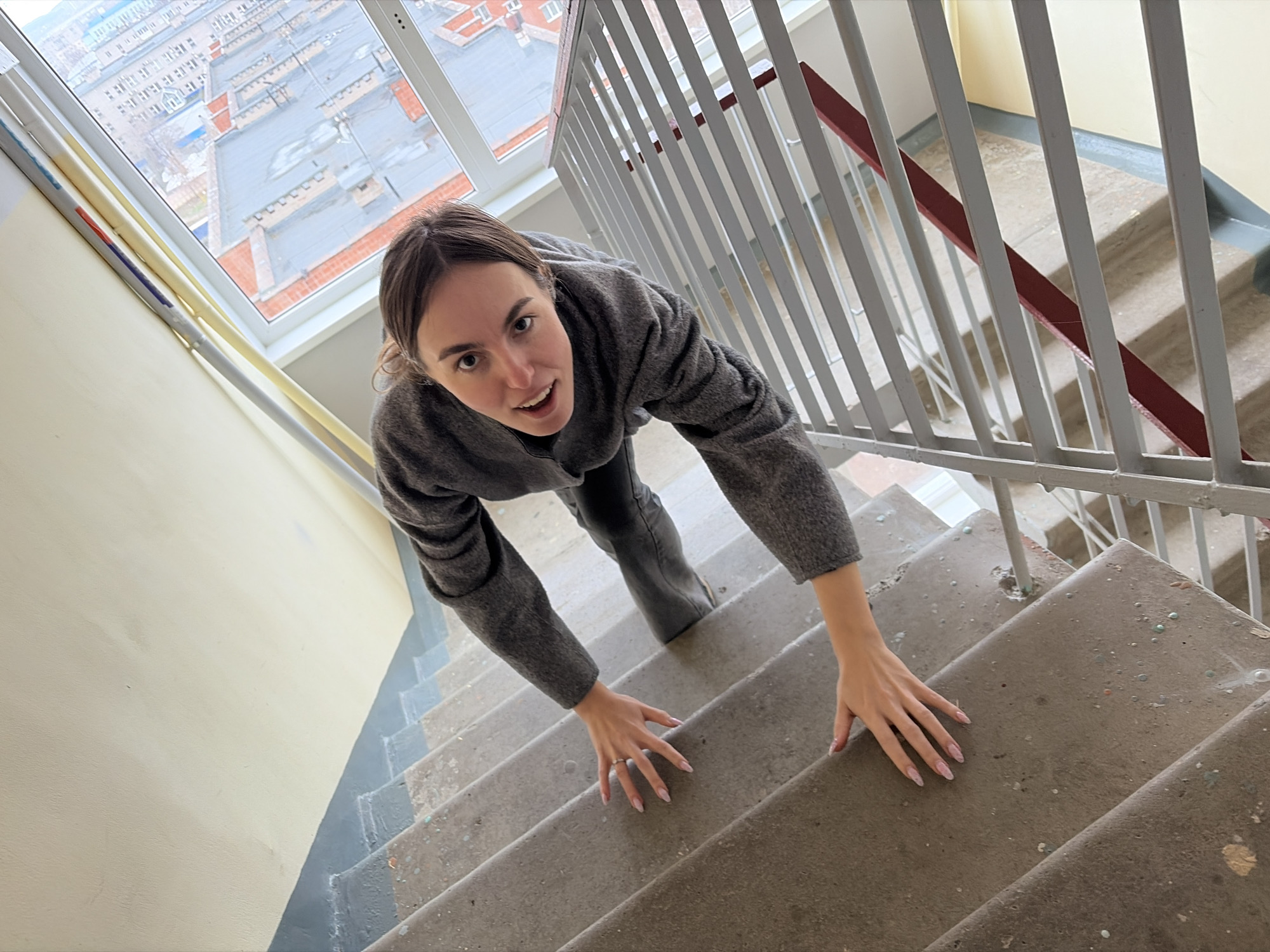 A young woman crawls up a concrete staircase in an indoor setting. She wears a gray sweater and jeans, and has manicured nails. The staircase is enclosed by a white metal railing with a red accent. A window reveals a cityscape, identified as Izhevsk, Russia. The surrounding walls are painted in a muted yellow color. The scene appears to be a stairwell in a building.
