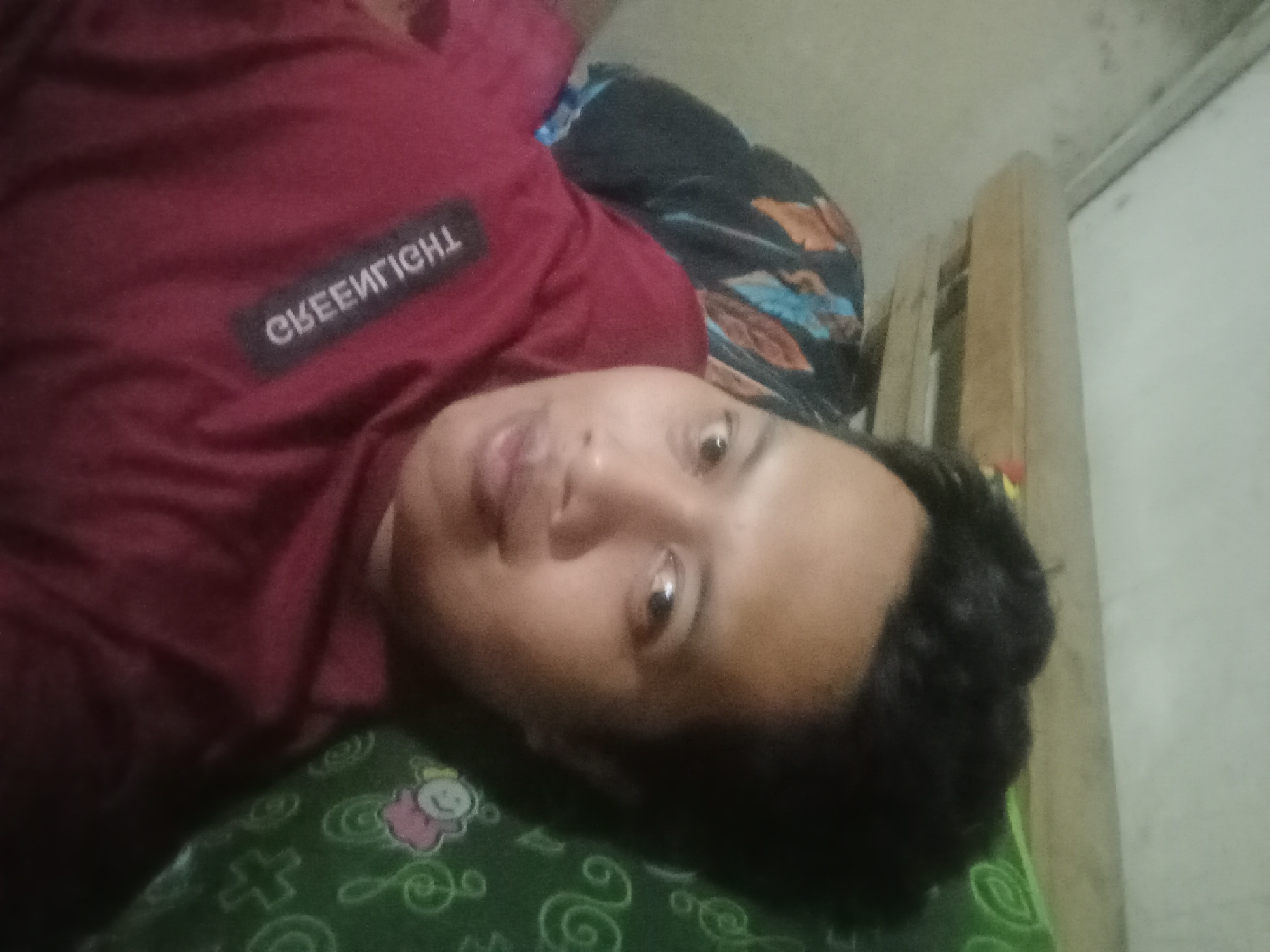 A selfie shows a person lying down, with their face partially visible. They are wearing a maroon shirt with a black rectangular patch that has white text. The text on the patch reads "CREENIGHT". The person's head is resting on a green patterned pillow or blanket. The background appears to be an indoor setting, possibly a bedroom, with a wooden structure and a wall visible. The lighting is dim, and the image is slightly blurry. There is no indication of the specific location in Cilegon, Indonesia, other than the overall context.