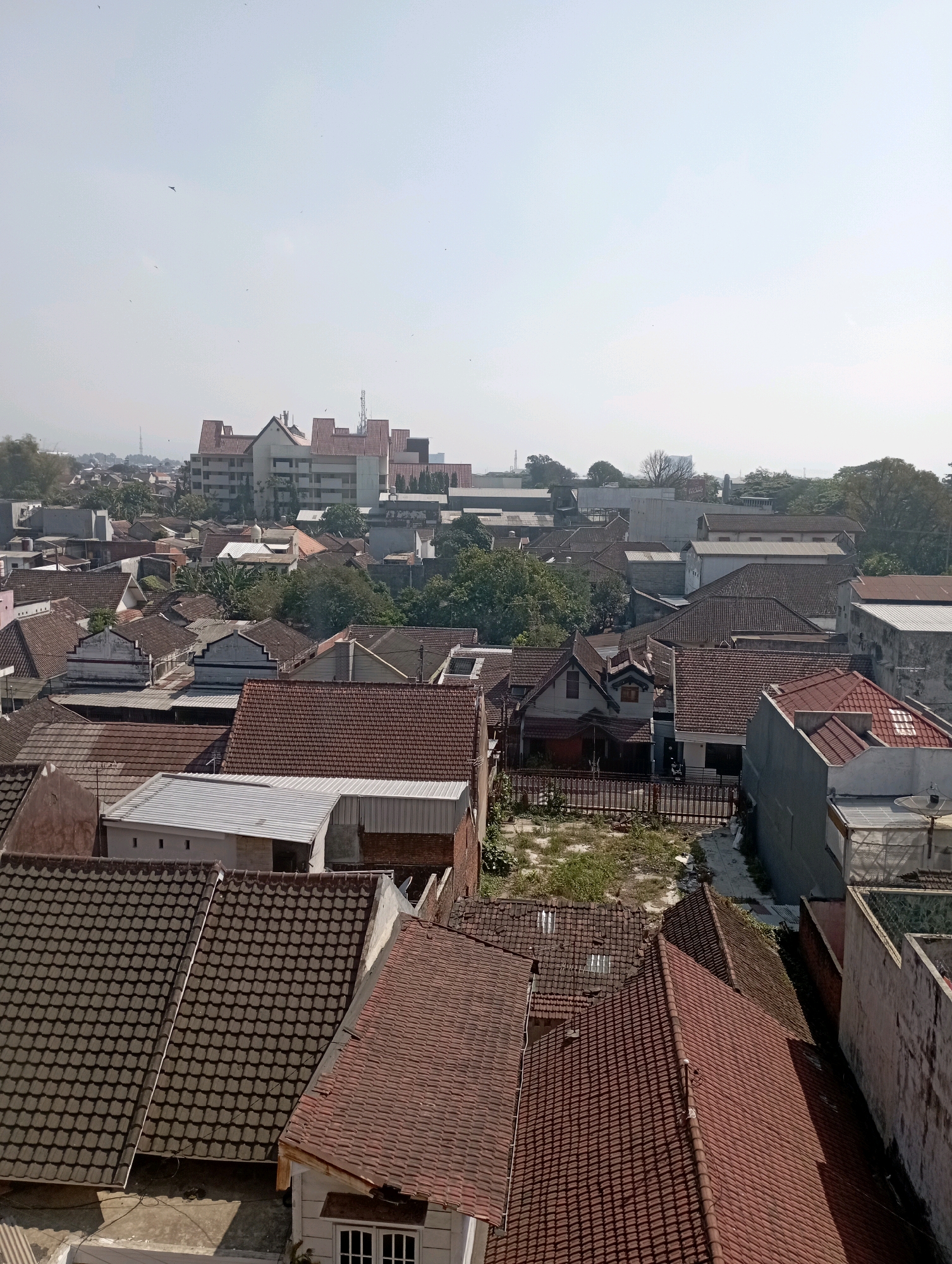 This aerial photograph showcases a dense urban landscape dominated by residential buildings with terracotta tile roofs. The structures vary in size and architectural style, with many featuring single or two-story designs. Some buildings exhibit white or light-colored walls, while others are made of brick. Greenery is interspersed throughout the scene, with trees visible between the houses, suggesting a moderate level of vegetation within the city. In the background, a larger, multi-story building with a distinct red-tiled roof and a light-colored facade stands out, possibly a commercial or institutional building. The sky is clear and bright, indicating a sunny day, and a few birds are visible in flight. The overall impression is of a well-established neighborhood in a tropical or subtropical region. Given the context provided, this scene is likely located in the city of Malang, Indonesia, a region known for its residential development and proximity to natural landscapes.