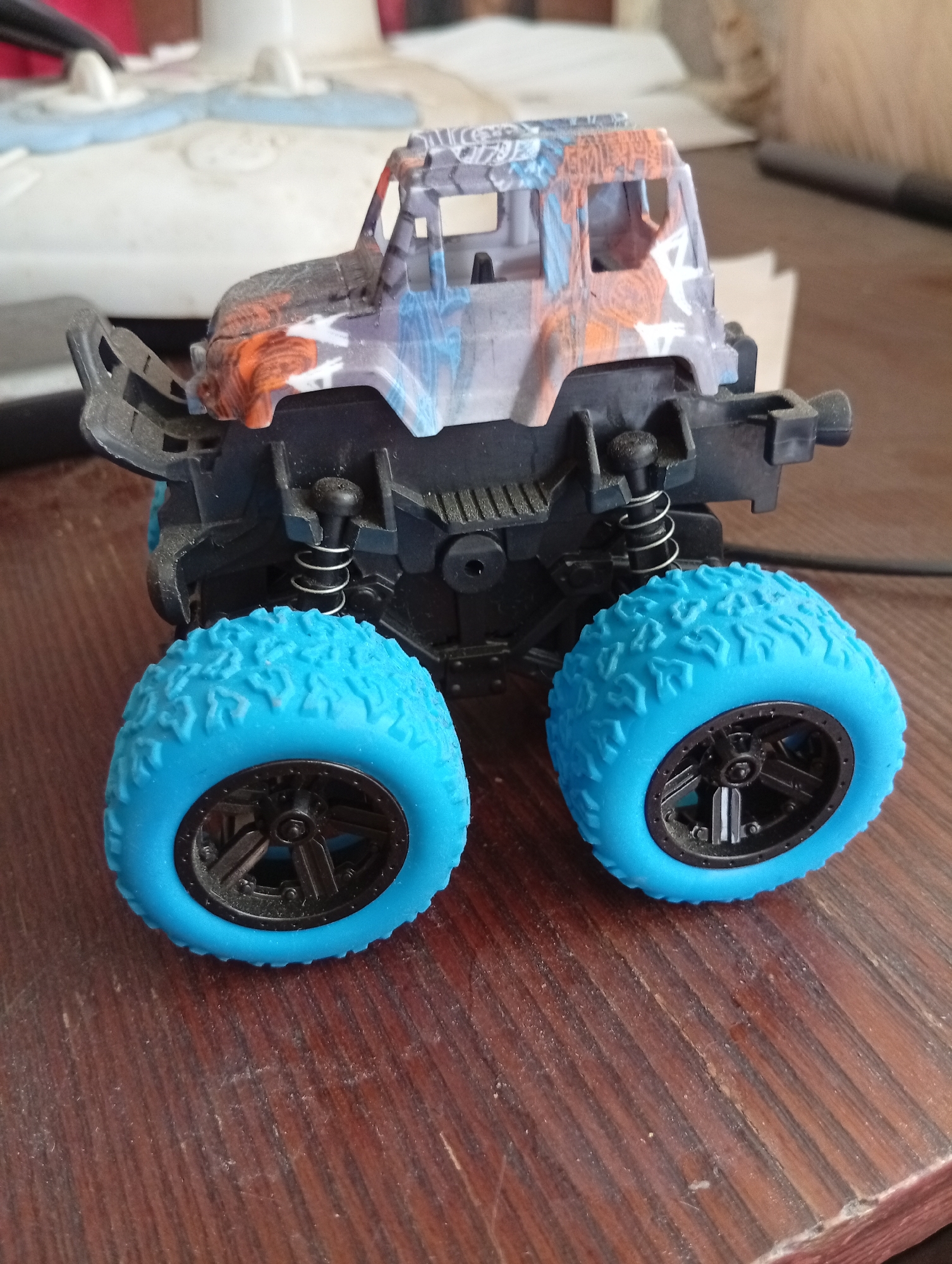 A close-up, high-angle shot captures a toy monster truck with a distressed paint job on a wooden surface. The truck's body features a camouflage pattern in shades of gray, blue, and orange. It has oversized, bright blue tires with intricate tread patterns and black rims. The suspension system, with exposed springs, is visible beneath the body. The wooden surface appears worn, with visible grain and some dust particles. The background is out of focus, showing a white object and possibly other household items. The image is an indoor shot. There is no indication of location in Kyshtym, Russia.