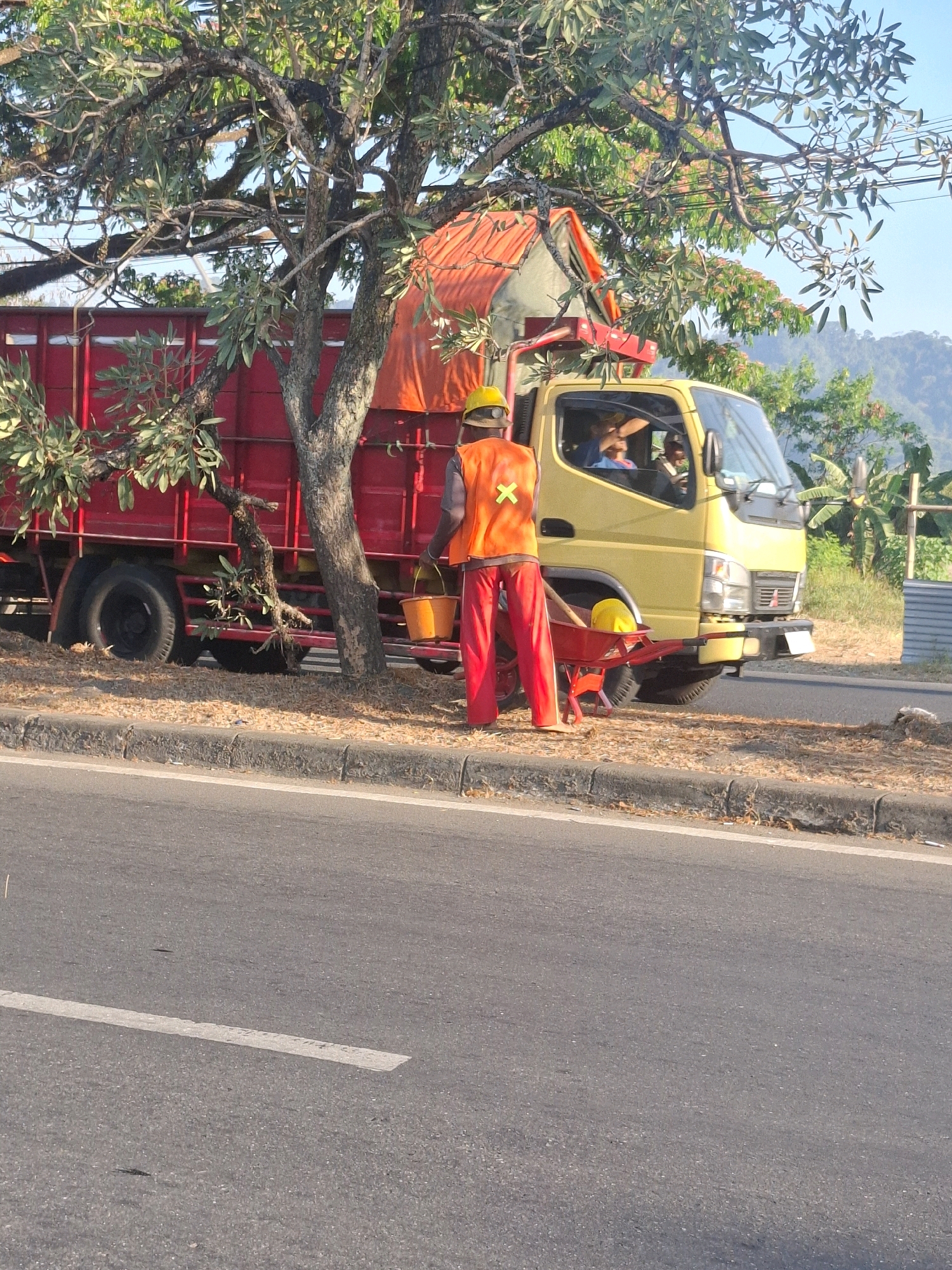 In the city of Sukabumi, Indonesia, a man wearing a yellow hard hat and an orange reflective vest stands beside a bright red truck. He is holding an orange bucket in his left hand and another identical bucket hangs from the back of a red wheelbarrow positioned to his right. He is wearing red pants and appears to be barefoot. Inside the yellow truck's cab, two individuals are visible. The truck has a red cargo bed with a tarp covering its contents. The scene is situated on the side of a paved road, with a concrete curb and a scattering of dry leaves on the ground. A large tree with dense foliage extends over the truck. In the background, a lush green landscape with rolling hills is observed under a clear blue sky.