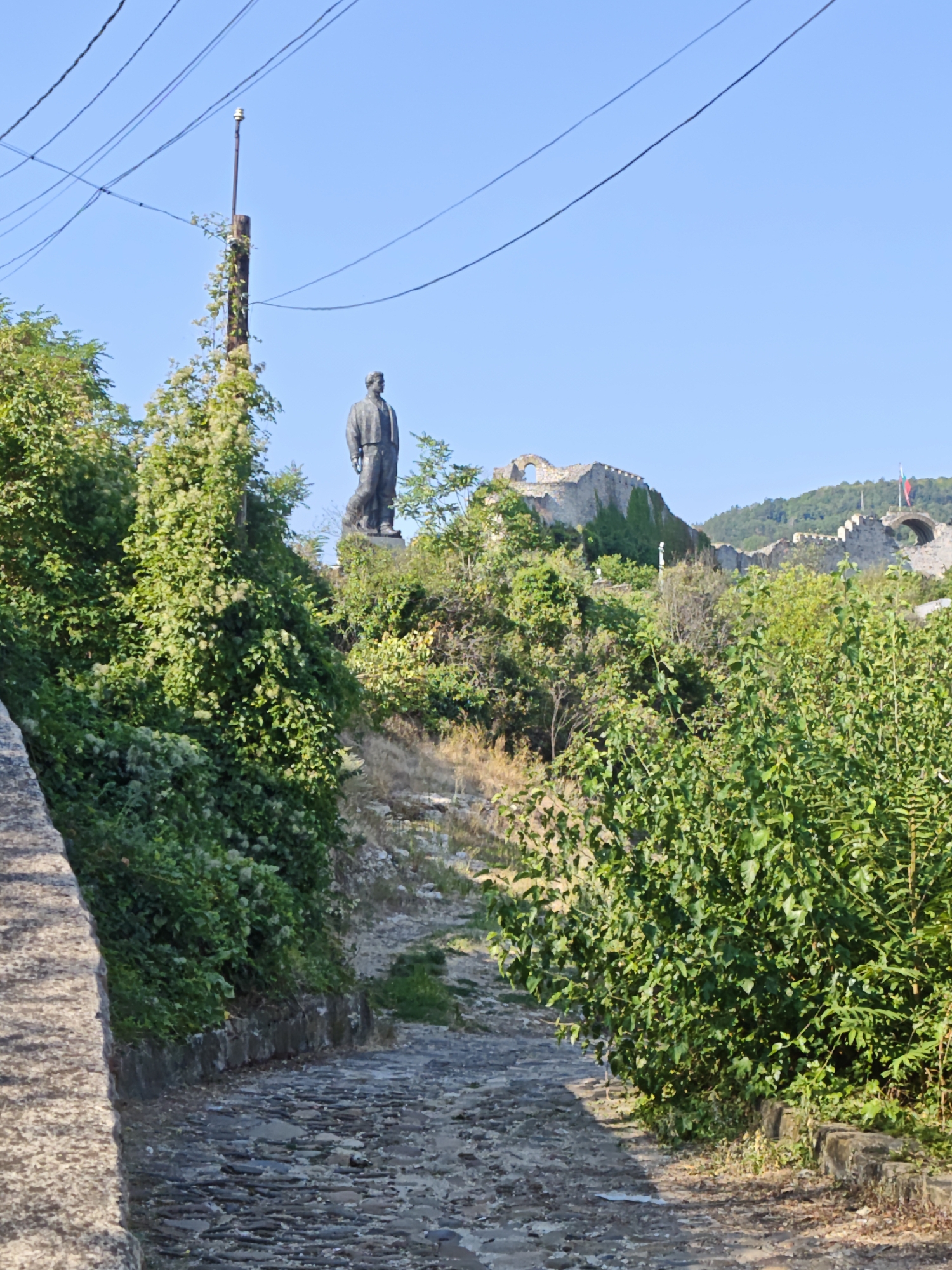 The image depicts a cobblestone path winding uphill through lush green foliage. In the background, a large bronze statue of a man stands prominently on a hill, overlooking the path. Beyond the statue, ancient stone ruins, possibly a fortress or castle, are visible, with ivy climbing up the weathered walls. The sky above is clear and blue, with a few power lines strung across it. The scene is set in the city of Lovech, Bulgaria, likely at or near the Varosha quarter, which is known for its historical architecture and the prominent statue. The overall impression is one of historical grandeur combined with the natural beauty of the surrounding landscape.