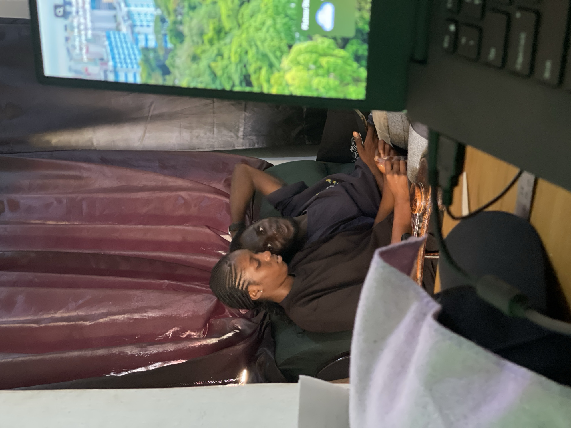 In Durumi, Nigeria, two young people are seen in a living room setting. A woman, with her hair in braided cornrows, is seated on a dark green couch, leaning against the backrest. She is wearing a dark brown, long-sleeved shirt. Beside her, a young man is also seated on the couch, resting his head on her shoulder. He is wearing a dark blue t-shirt. The woman is looking down at a smartphone she is holding in her hands, while the man is looking towards the viewer. A laptop is positioned in the foreground, partially obscuring the scene. The laptop screen displays a vibrant green landscape with buildings in the distance. Behind the couch, a dark, textured curtain is visible, with a lighter colored wall to the left. On a table to the left of the couch, a white cloth is draped over a dark object, possibly a laptop bag or a cushion. A mouse and a wire are also visible on the table. The overall atmosphere suggests a casual, relaxed moment.
