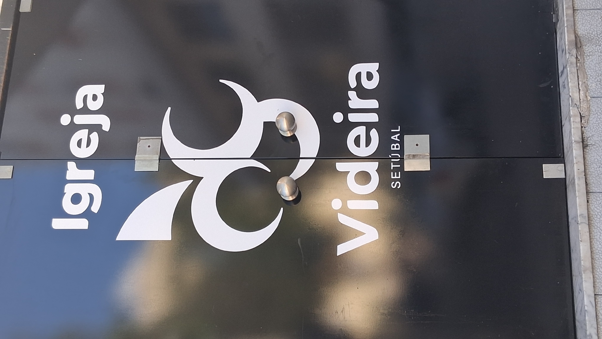 A black door has white lettering and a logo on it. The lettering reads "Igreja" at the top and "Videira" below a logo in the middle. Underneath "Videira" is the word "SETÚBAL". The logo appears to be a stylized representation of the letters 'AV' or similar shapes that form abstract swirls. Two metallic door handles are visible in the center of the door, positioned between the logo and the word "Videira". The door is divided vertically in the middle, and there are metallic fixtures on the door at various points. The text and logo are illuminated, suggesting they are on an exterior surface. The word "SETÚBAL" indicates the location of this establishment is in Setúbal, Portugal.