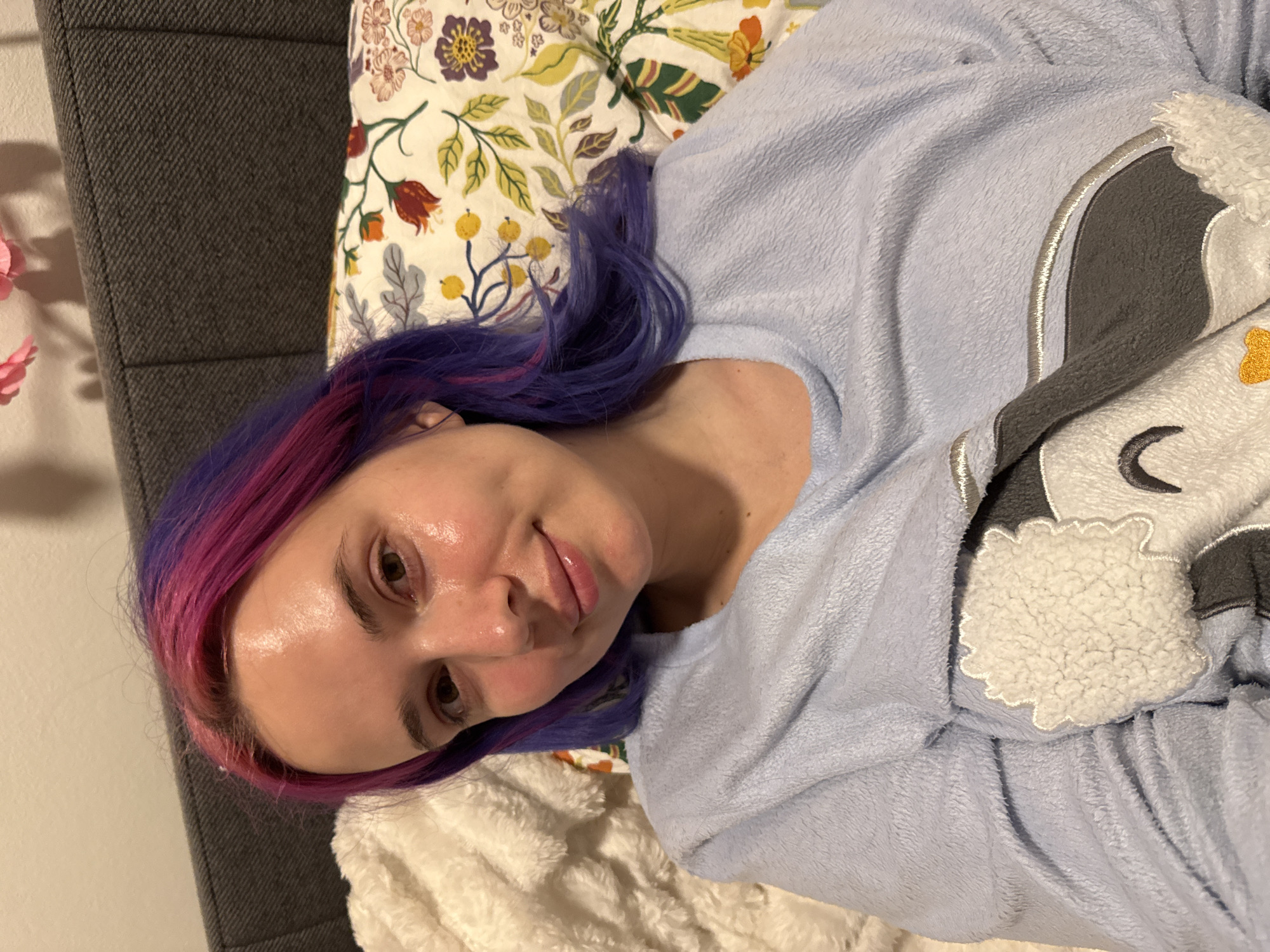 A woman with pink and purple hair is lying on a bed. She is wearing a light blue sweater with a gray and white animal applique. The bed has a floral patterned sheet and a white fuzzy blanket. A gray headboard is visible on the left side of the image. The photograph provides no information to ascertain its location.