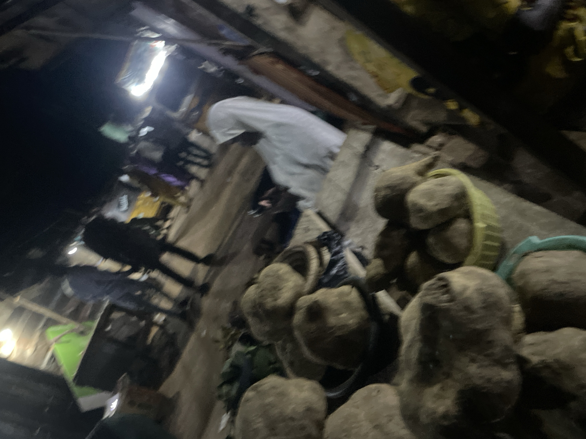 This is a nighttime outdoor scene, likely in a market, possibly in Lokoja, Nigeria. The image is blurry and dark, with artificial lighting present. In the foreground, there are large, round, brown objects, possibly tubers or root vegetables, some of which are in containers. In the background, several people are visible, some appearing to be vendors and customers. One person is wearing a white garment, possibly a hijab. Structures resembling market stalls or shelters are present, with some covered in green material. The ground appears to be dirt or pavement. The overall impression is a busy market environment after dark.