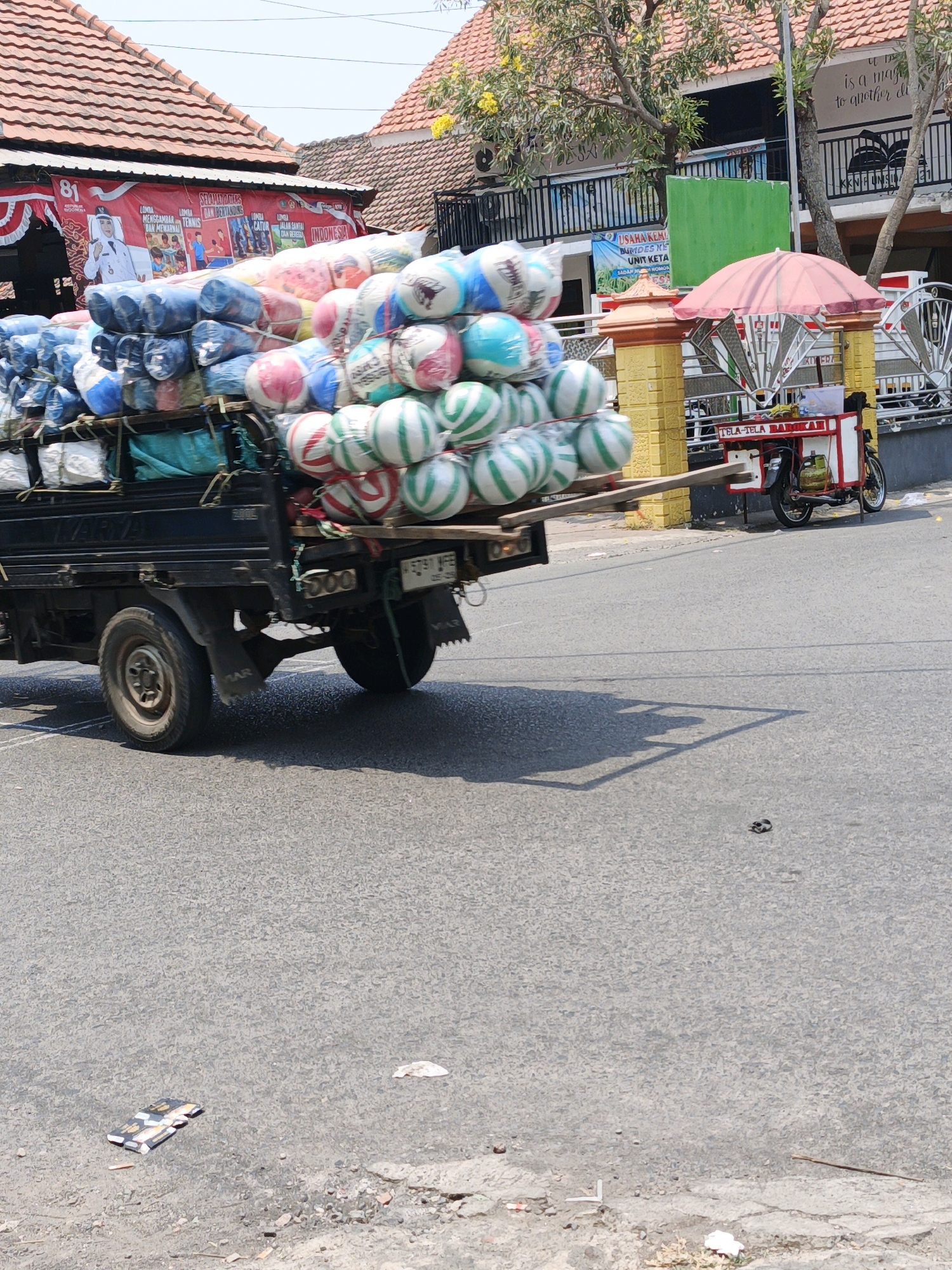 The image captures a truck overloaded with bundled goods on a street in Ketegan, Indonesia. The truck, a dark color with visible wheels and suspension, is laden with numerous round objects, some wrapped in plastic and others in striped patterns of green, white, and red. A large banner, featuring Indonesian text and images of people, is visible on a building behind the truck, suggesting a celebratory event or local advertisement. In the background, a pink umbrella shades a small cart, likely a vendor's stall, with a motorcycle parked nearby. The street itself is paved and appears to be an urban or semi-urban setting, with buildings and trees lining the road. The overall scene depicts a typical day with local commerce and transportation.