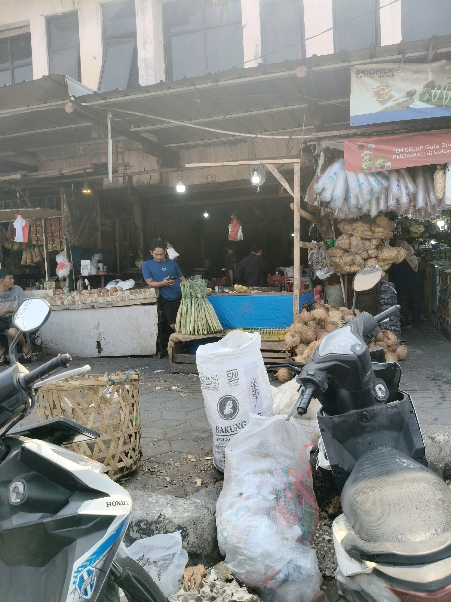 The image depicts an outdoor market scene in South Tangerang, Indonesia, characterized by a bustling atmosphere with various stalls and goods. In the foreground, two motorcycles are parked on a paved area. One is a white and blue Honda scooter, partially visible on the left, and the other is a black scooter on the right, with its seat facing the viewer.
Several large woven baskets are scattered around, one of which is positioned in front of the white and blue scooter. Next to the black scooter, a large white sack is prominent, displaying "HALAL" and "SNI" logos, suggesting it contains food items. Another large, clear plastic bag filled with miscellaneous items is also near the scooters.
The background reveals a market stall with goods displayed on shelves and hanging from the ceiling. A man in a blue t-shirt is standing near a stall, looking at his phone. Another person is seated on the left, wearing a grey t-shirt. Various produce, including coconuts and what appears to be bundles of leeks or similar vegetables, are piled up. Banners with Indonesian text are visible above the stalls, one featuring an "INDOMILK" advertisement and another promoting "TEH CELUP GULA 2 IN 1". The overall environment suggests a typical day at a local Indonesian market.