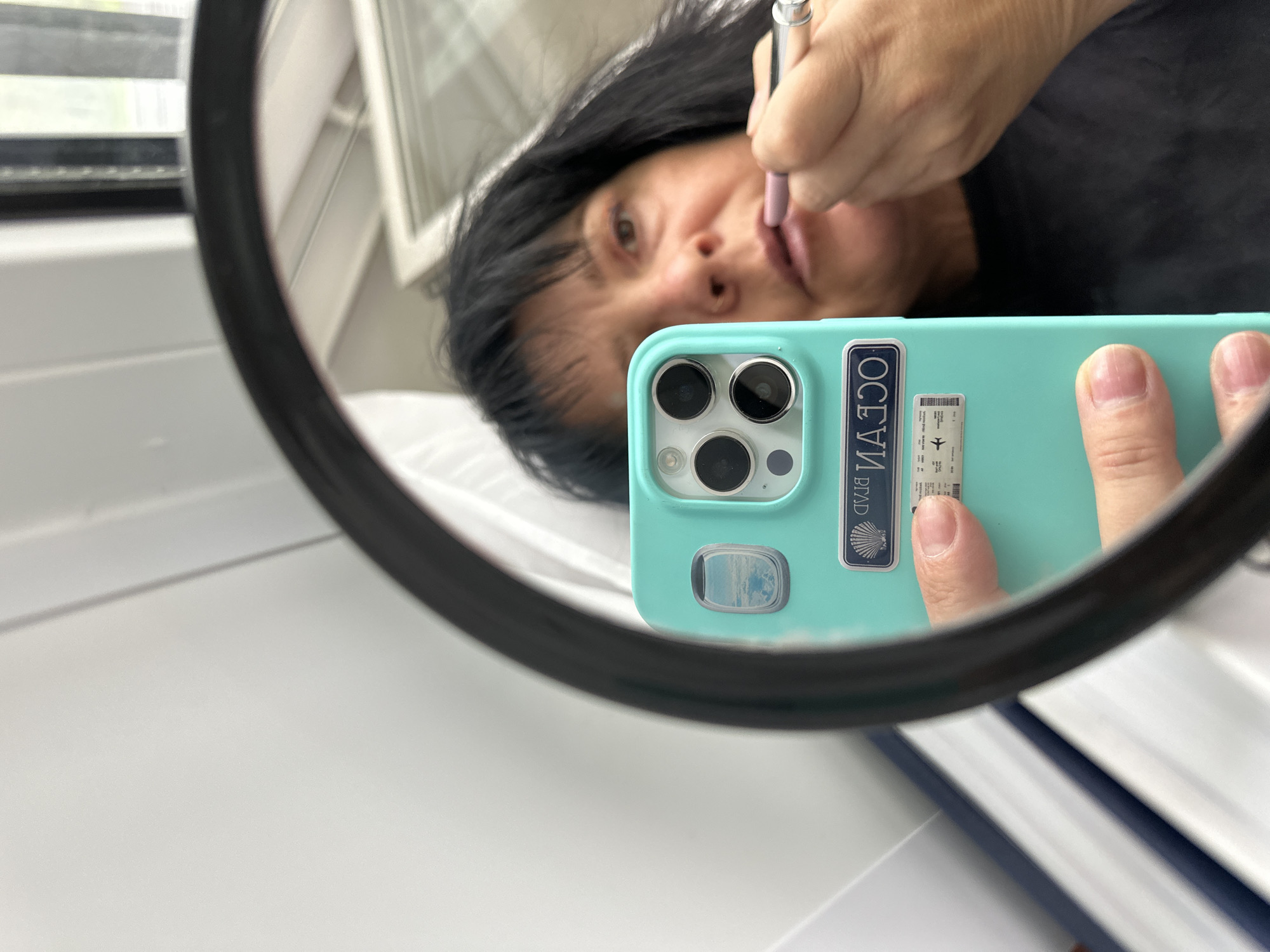 A woman is taking a selfie in a mirror, applying lipstick with a makeup pencil. She is holding a light blue phone with stickers on the back, one a rectangular “Ocean Beach” sticker and another that looks like an airline boarding pass. The mirror appears to be round and is placed against a white surface. The environment is indoors, possibly a bathroom or a similar space with white walls and a window in the background. There is no indication of the location being in Dnp Ryzhiki, Russia.