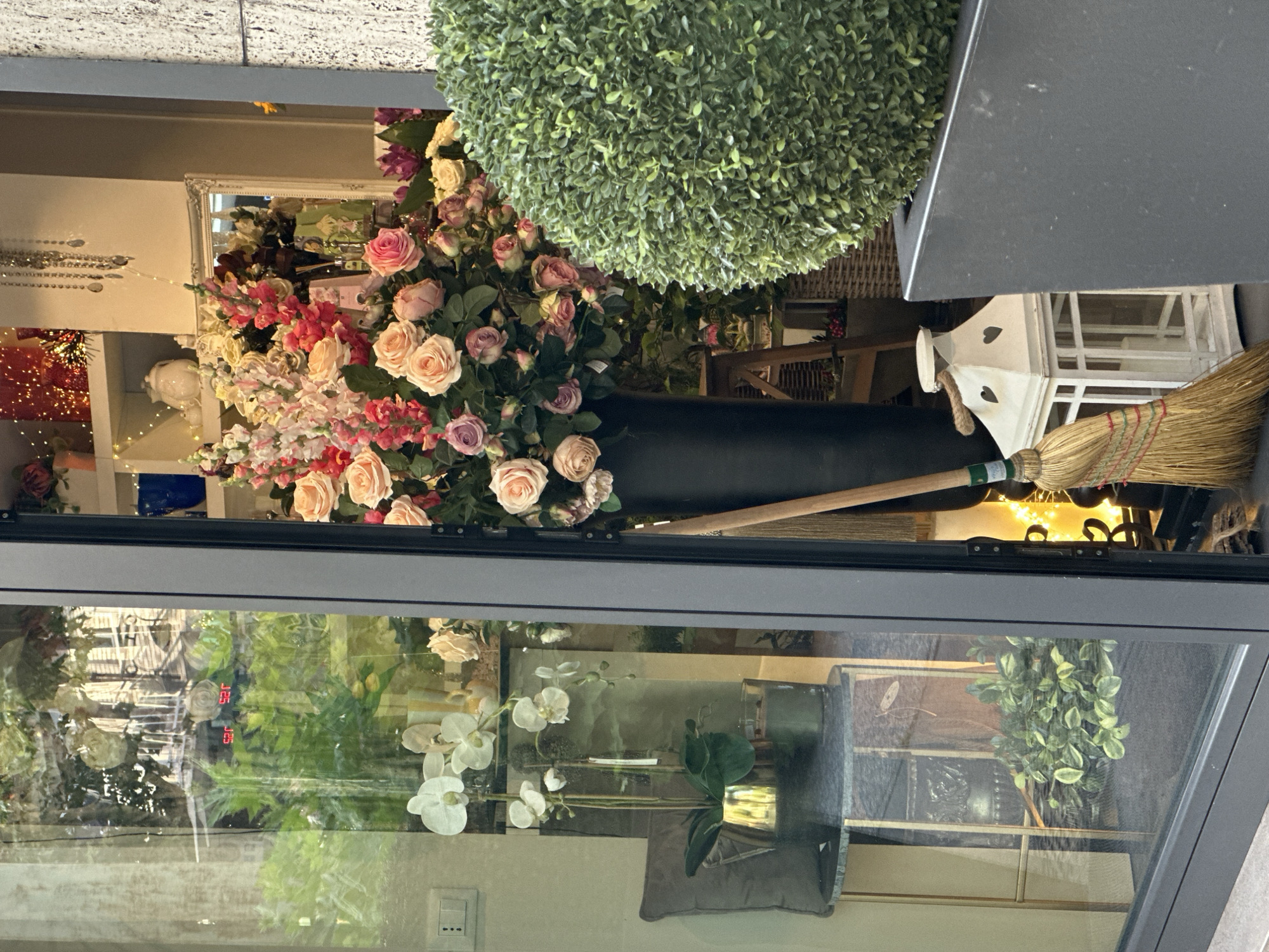 A floristry shop window in Trieste, Italy, displays a large bouquet of pink and cream roses and snapdragons, a tall black vase, a white lantern with heart cutouts, and a broom. To the left, through the glass, a white orchid plant in a gold pot sits on a marble-topped stand next to a candle. Behind the orchid, a digital thermometer displays 25.2 degrees Celsius and 25 degrees Fahrenheit. The exterior facade of a building with textured stonework is visible to the right.