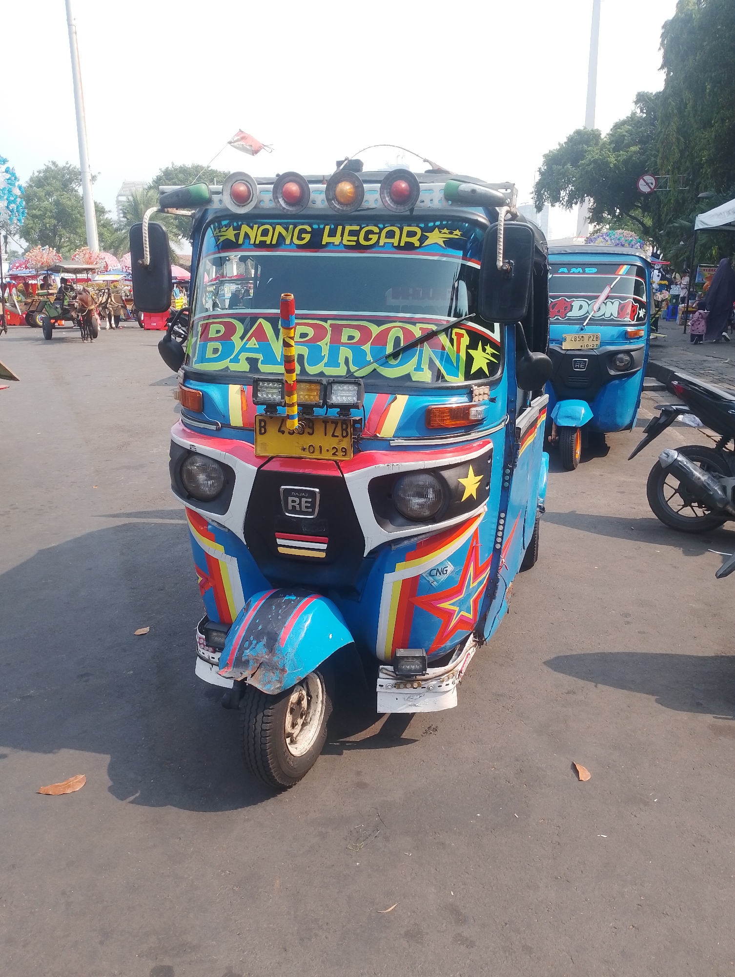 In Central Jakarta, Indonesia, a vibrant blue and colorful auto-rickshaw, commonly known as a "Bemo," is parked on an asphalt road. The vehicle is heavily decorated with an array of stickers and paintings, including the name "BARRON" in large, stylized yellow letters across the windshield. Additional colorful graphics, such as red and yellow stripes and yellow stars, adorn the front and sides of the auto-rickshaw. It has multiple lights on its roof, some of which are illuminated. A license plate is visible, reading "B 4593 TZB" and "01-29." The auto-rickshaw has a single front wheel and two rear wheels. Another auto-rickshaw, also blue and decorated, is visible behind the primary subject, along with a black motorcycle parked to the right. In the background, trees and a tall white pole are present, suggesting an outdoor, possibly urban, environment. A few fallen leaves are scattered on the road. The scene appears to be captured during daylight.