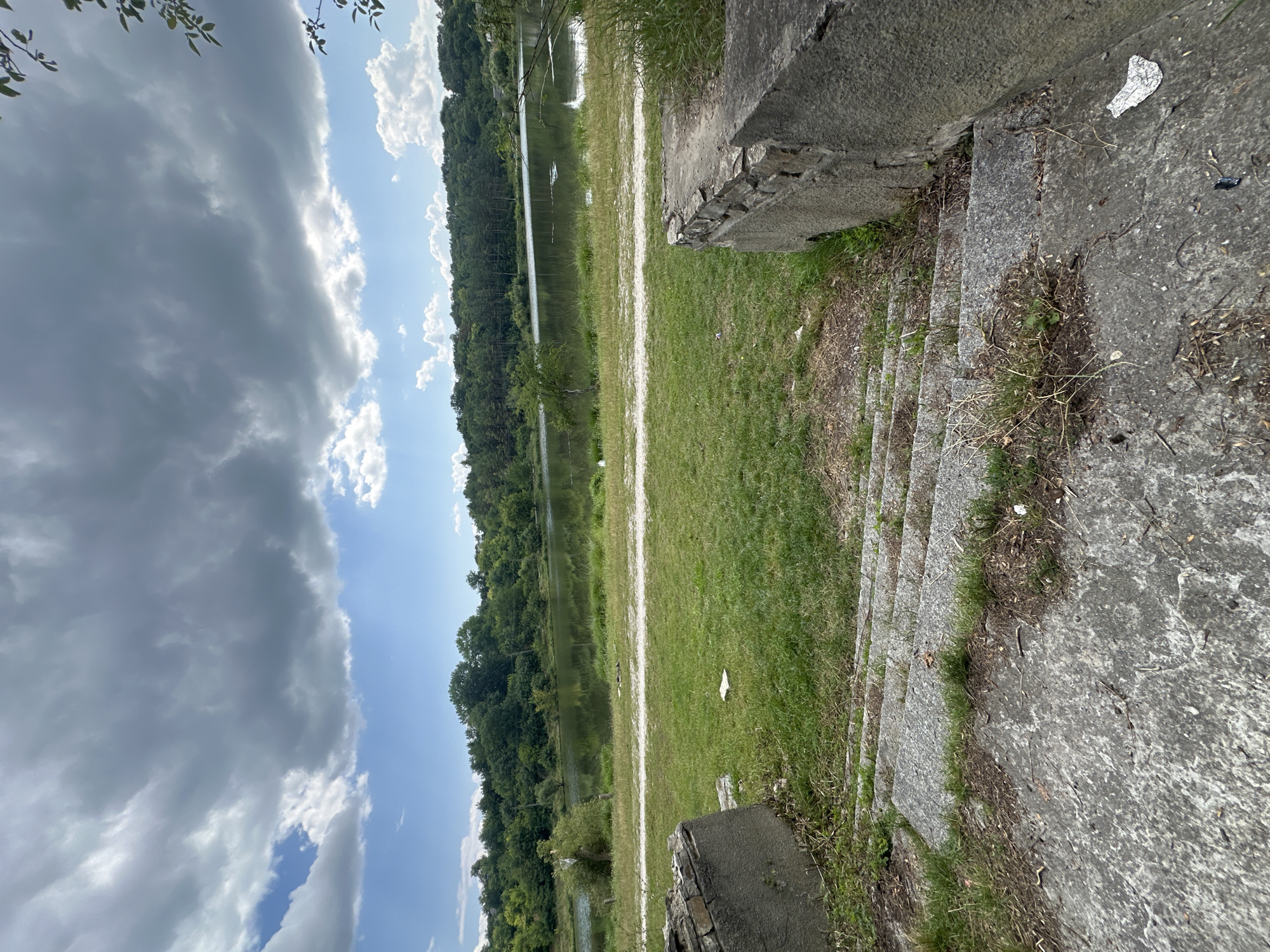 The image depicts a view of a lake surrounded by trees, under a cloudy sky. In the foreground, there are weathered concrete steps leading down from an elevated grassy area. On the left and right sides of the steps, there are also remnants of concrete structures, possibly part of a building or barrier. The grassy area shows some scattered debris. The lake is calm and reflects the greenery of the trees and the sky. The overall scene suggests an undeveloped or abandoned area near a body of water. This location is Makiv, Ukraine.