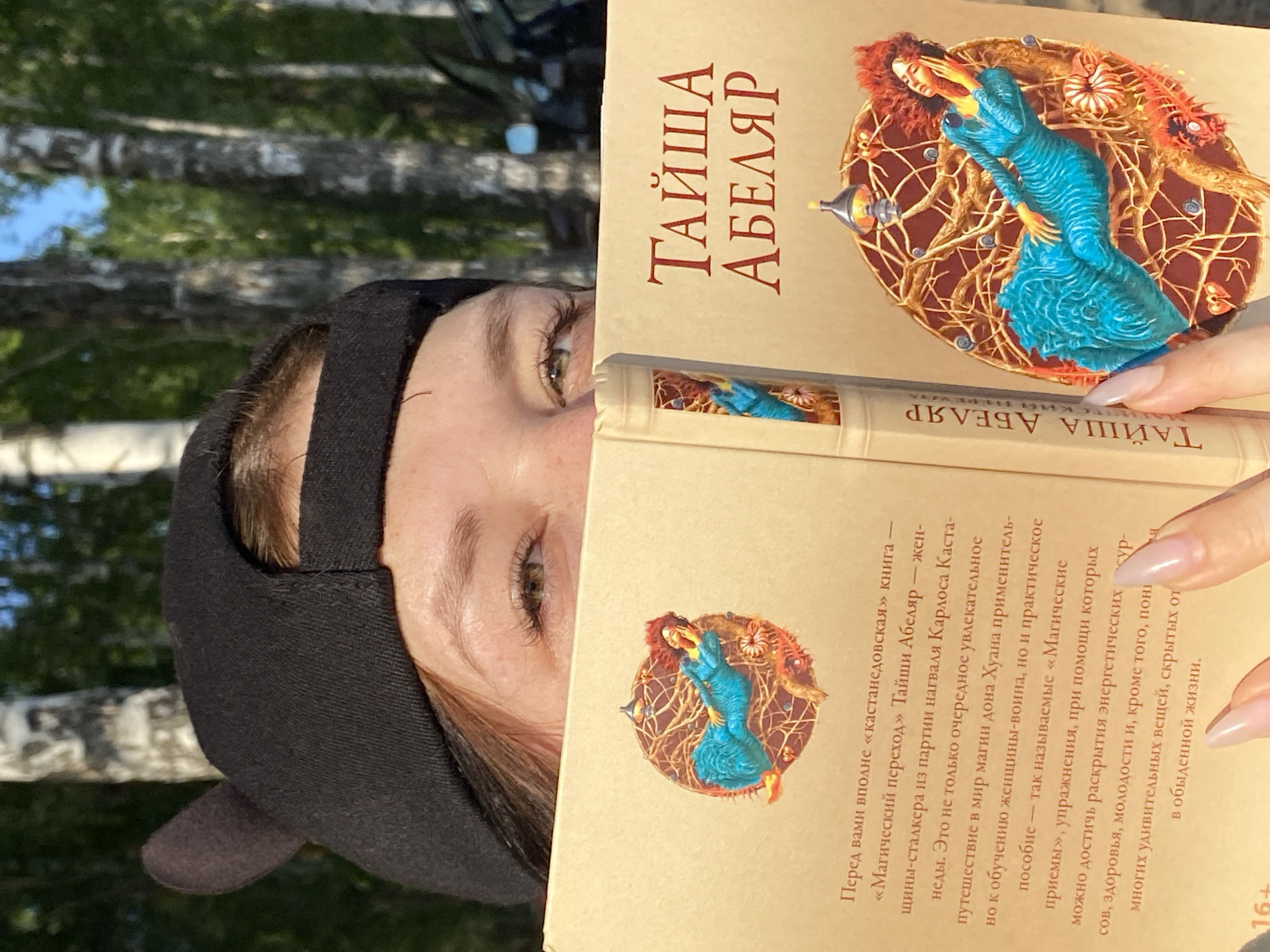 A woman in a black cap is holding a book titled "Taisha Abelyar" in Russian. The book's cover features an illustration of a woman in a blue dress surrounded by abstract designs. The woman holding the book has fair skin and is looking at the camera. The background of the image shows a blurred view of trees and greenery. The location is Noviy, Russia.