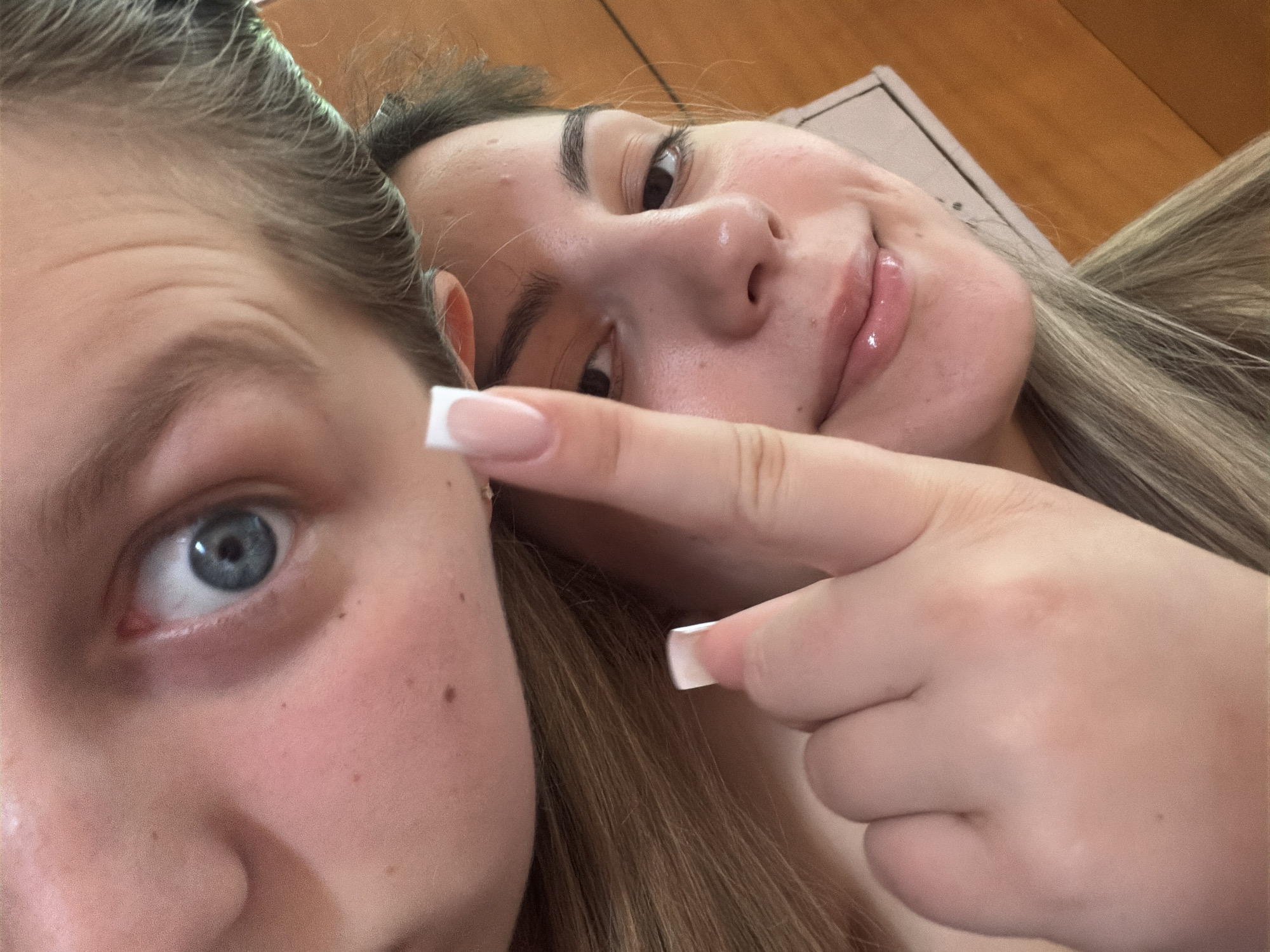 The image shows a close-up selfie of two young women. The woman in the foreground is on the right, with her face tilted upwards and a finger held up in front of her nose. She has dark hair, dark eyes, and appears to be smiling. She is wearing light-colored lip gloss. The woman in the background is on the left, and only her eye and forehead are visible. Her eye is open wide, and she has light-colored hair. Both women have long, blonde hair. The background appears to be an indoor setting with wooden furniture. There is no visible indication of the location being in Burgas, Bulgaria.