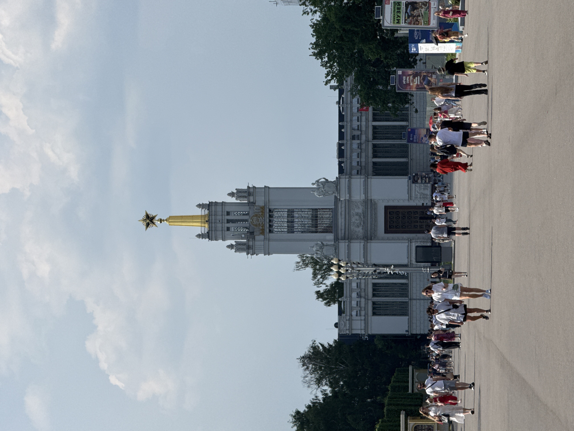 The image depicts the Pavilion of Agriculture, a prominent structure at the All-Russian Exhibition Centre (VDNKh) in Moscow, Russia. The pavilion is characterized by its imposing neoclassical architecture, featuring a central tower topped with a golden star. In front of the pavilion, a wide paved pathway is bustling with people strolling in various directions under a partly cloudy sky. The visitors are dressed in casual summer attire, suggesting a warm day. The scene is characterized by a sense of public gathering and leisure activity in an open-air urban environment.