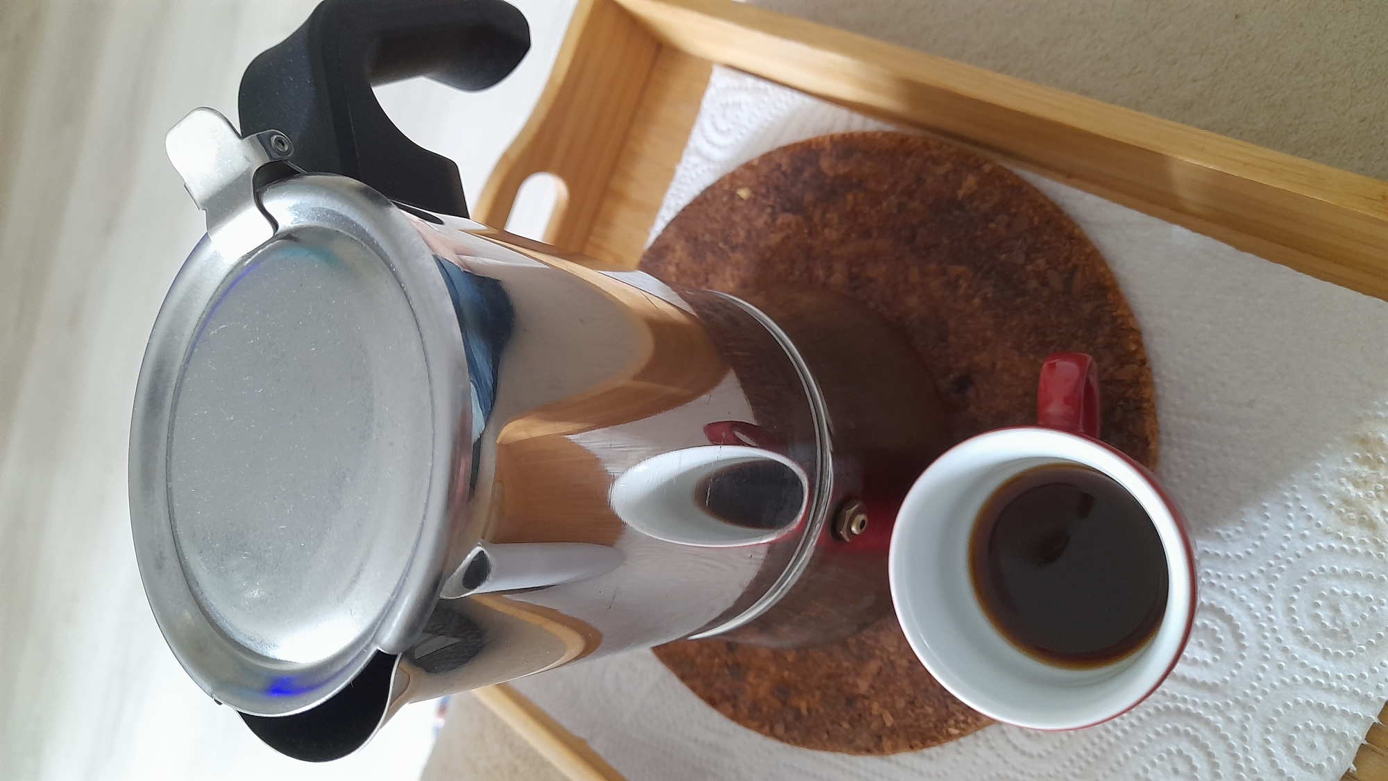 A Moka pot and a red mug containing coffee are arranged on a round cork coaster on a white napkin, which rests inside a wooden tray. The scene is brightly lit. The setting is unknown, and it is not possible to determine if the photo was taken in Saint Petersburg, Russia.
